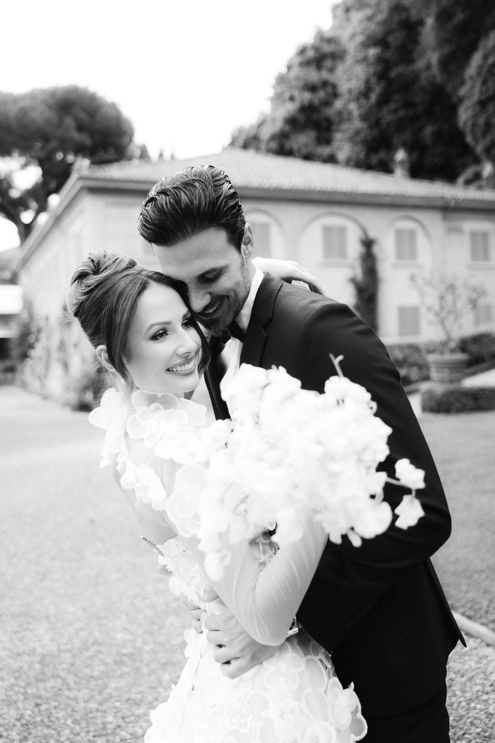Smiling bride holding white floral bouquet embraced by groom in black suit at Italian villa wedding.