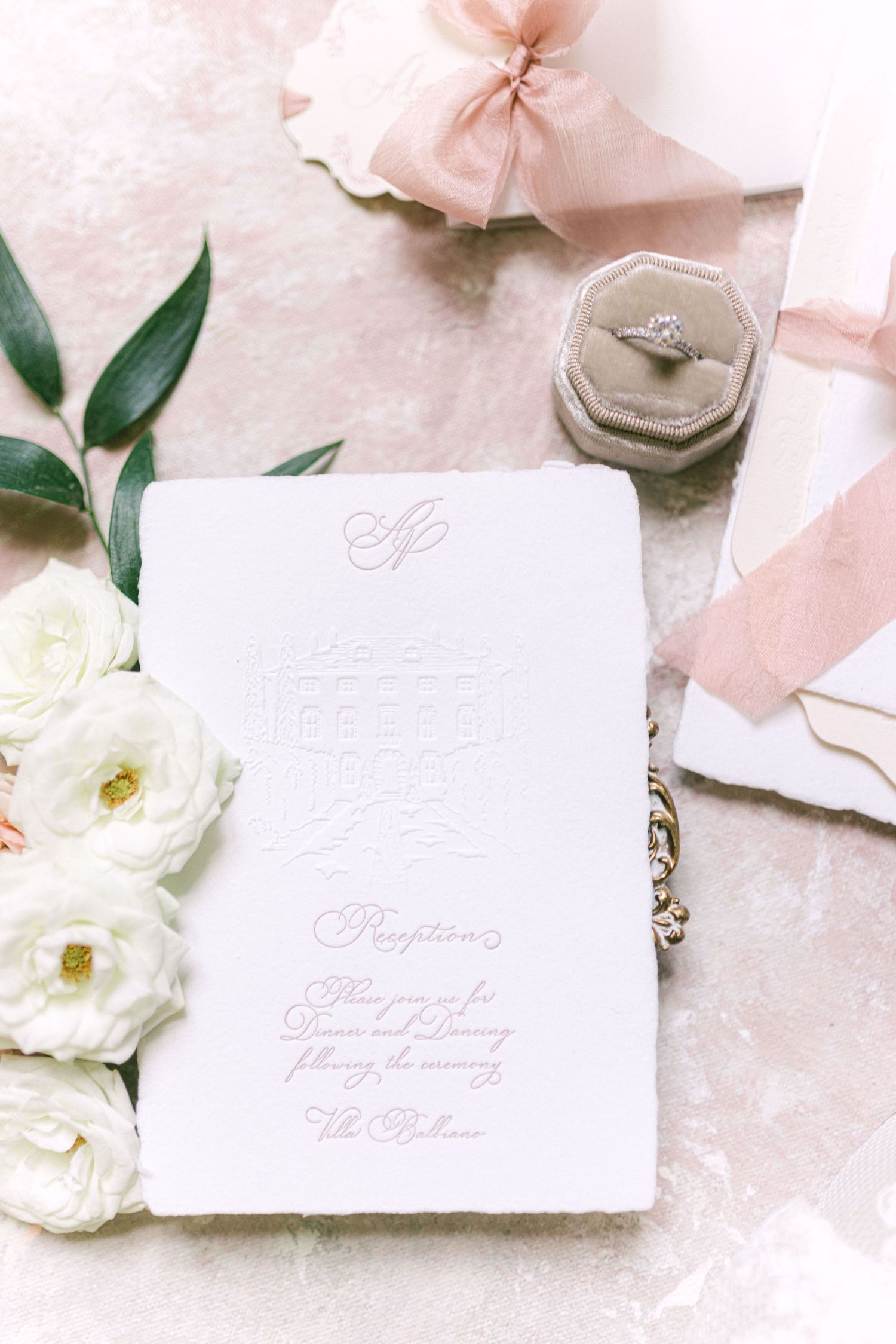 Wedding stationery flat lay with embossed invitation, diamond engagement ring in velvet box, white roses, and blush ribbon.