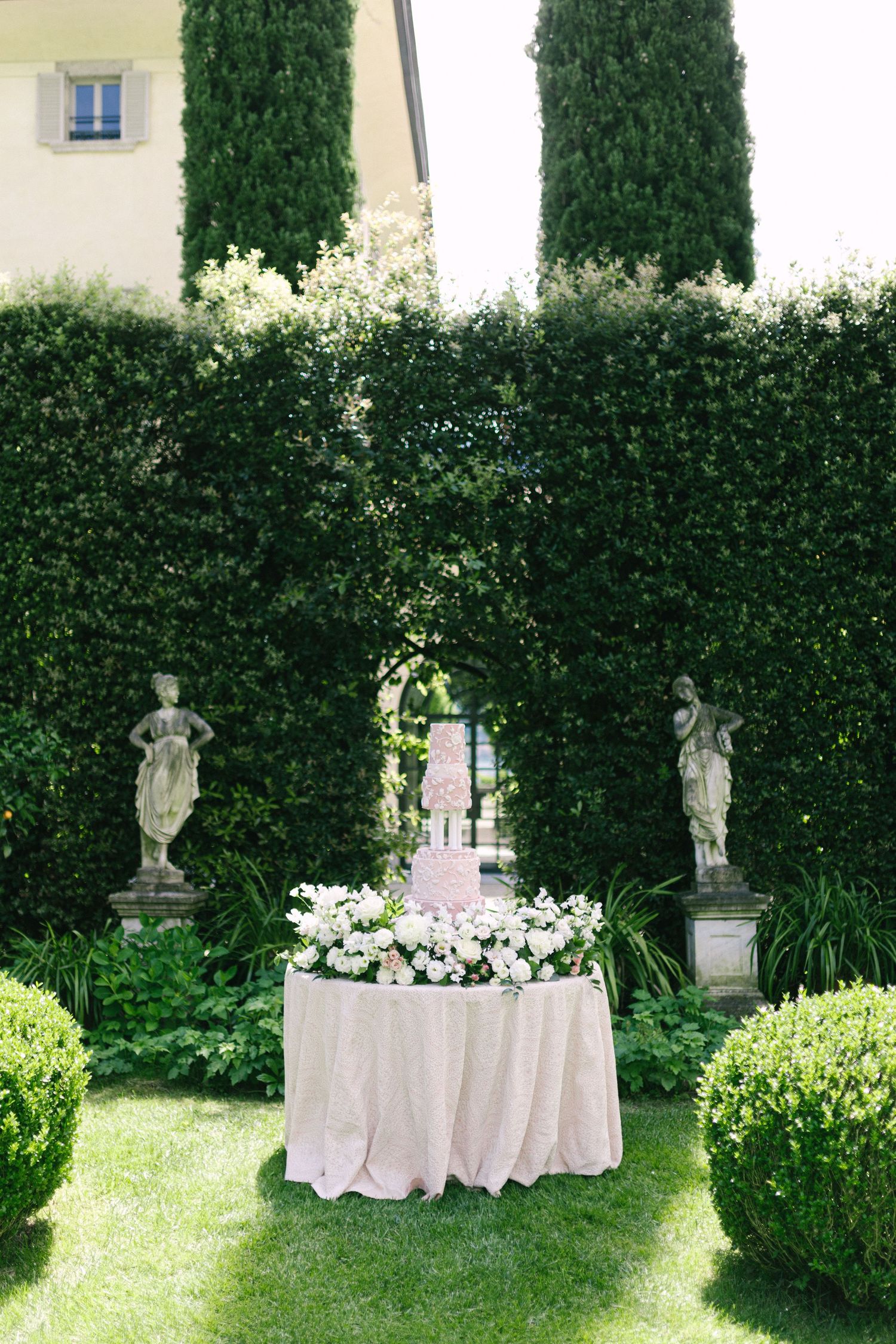 Elegant outdoor wedding cake display on draped table surrounded by white flowers, stone statues, and manicured hedges.