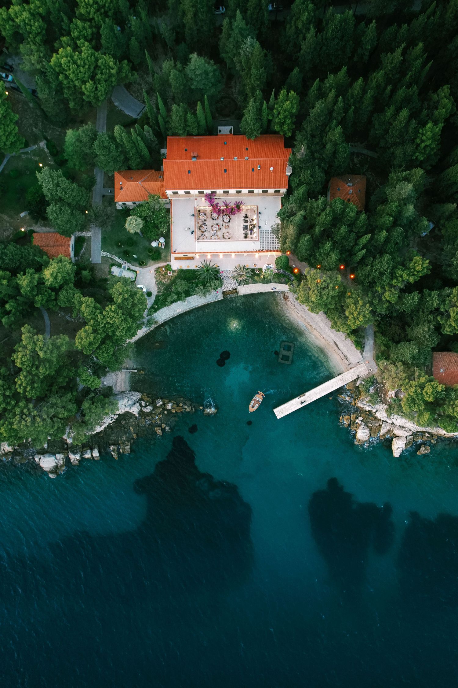 Aerial view of a waterfront villa with orange roof surrounded by lush green forest and turquoise water.