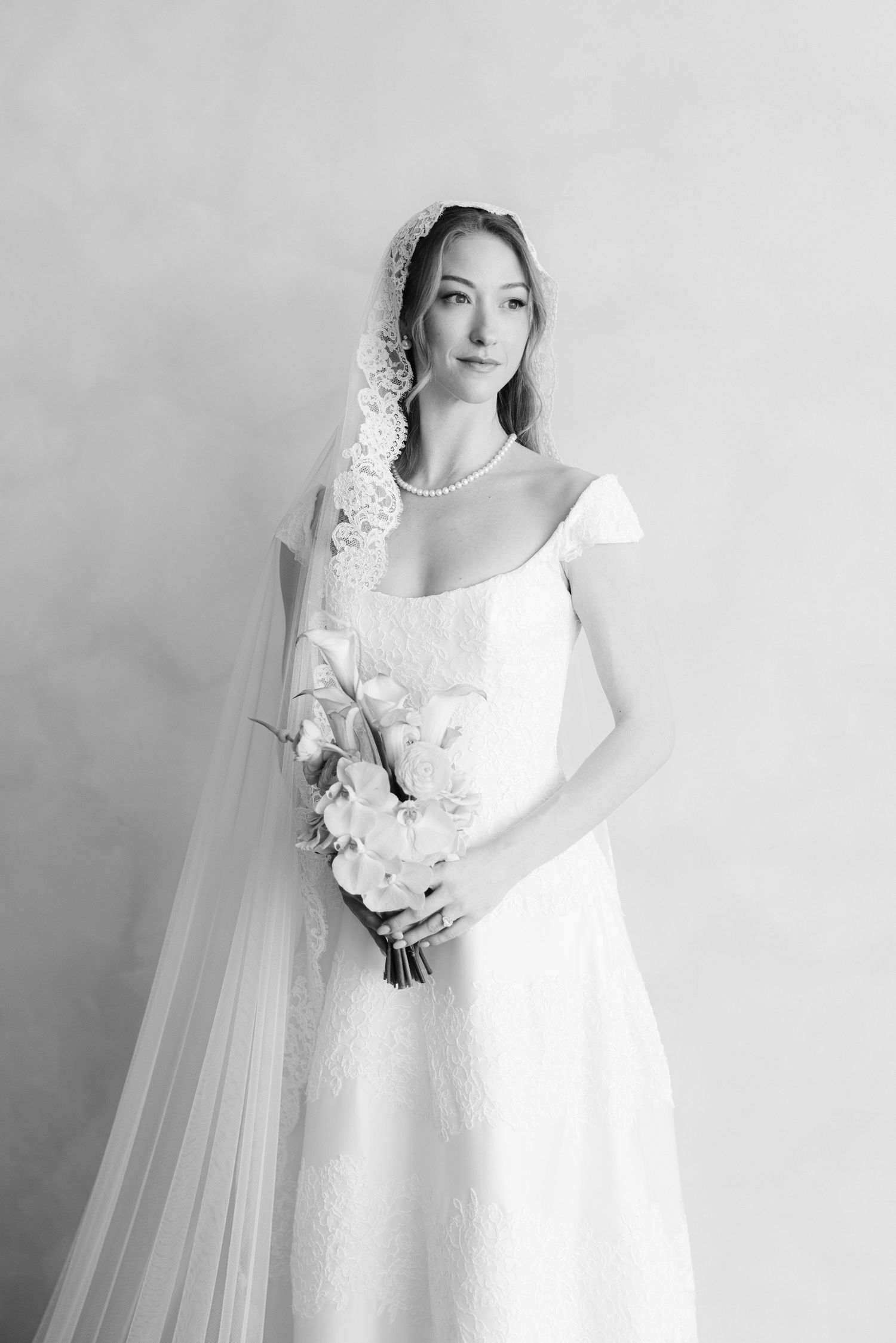 Black and white portrait of a bride in lace gown, pearl necklace, floral veil, holding a white bouquet.