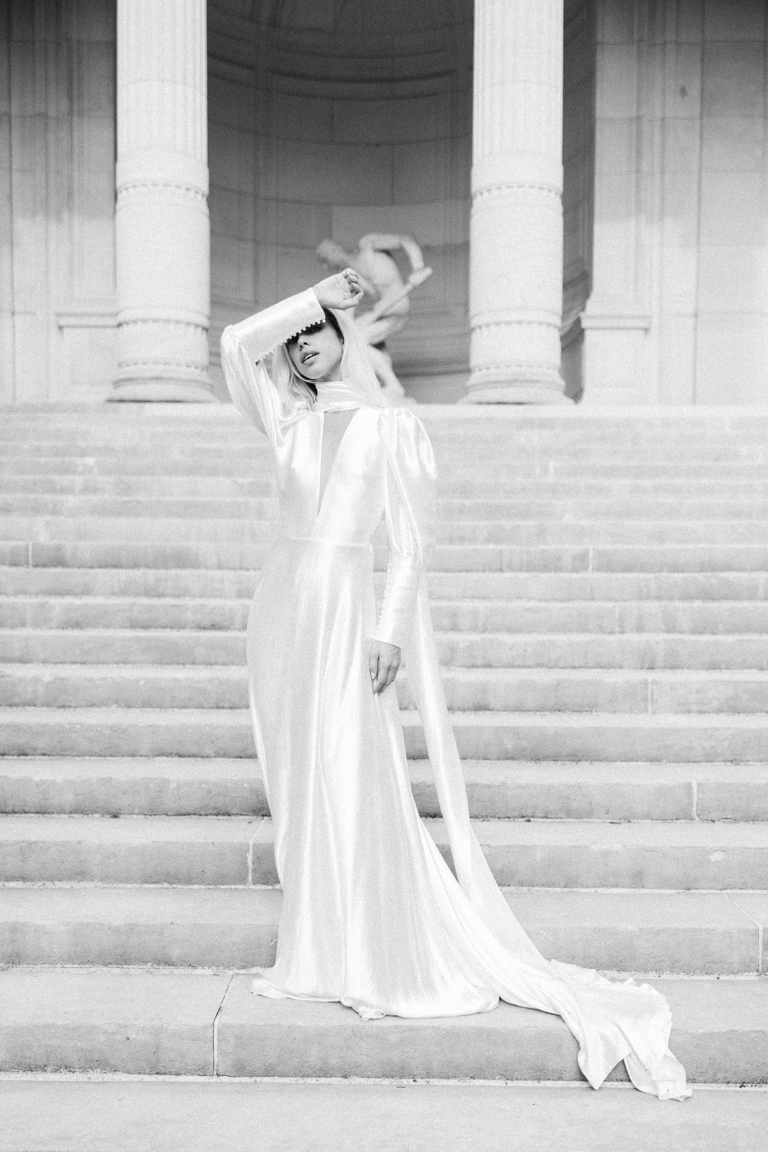 Bride in flowing white satin gown with long veil poses dramatically on grand stone staircase with columns.