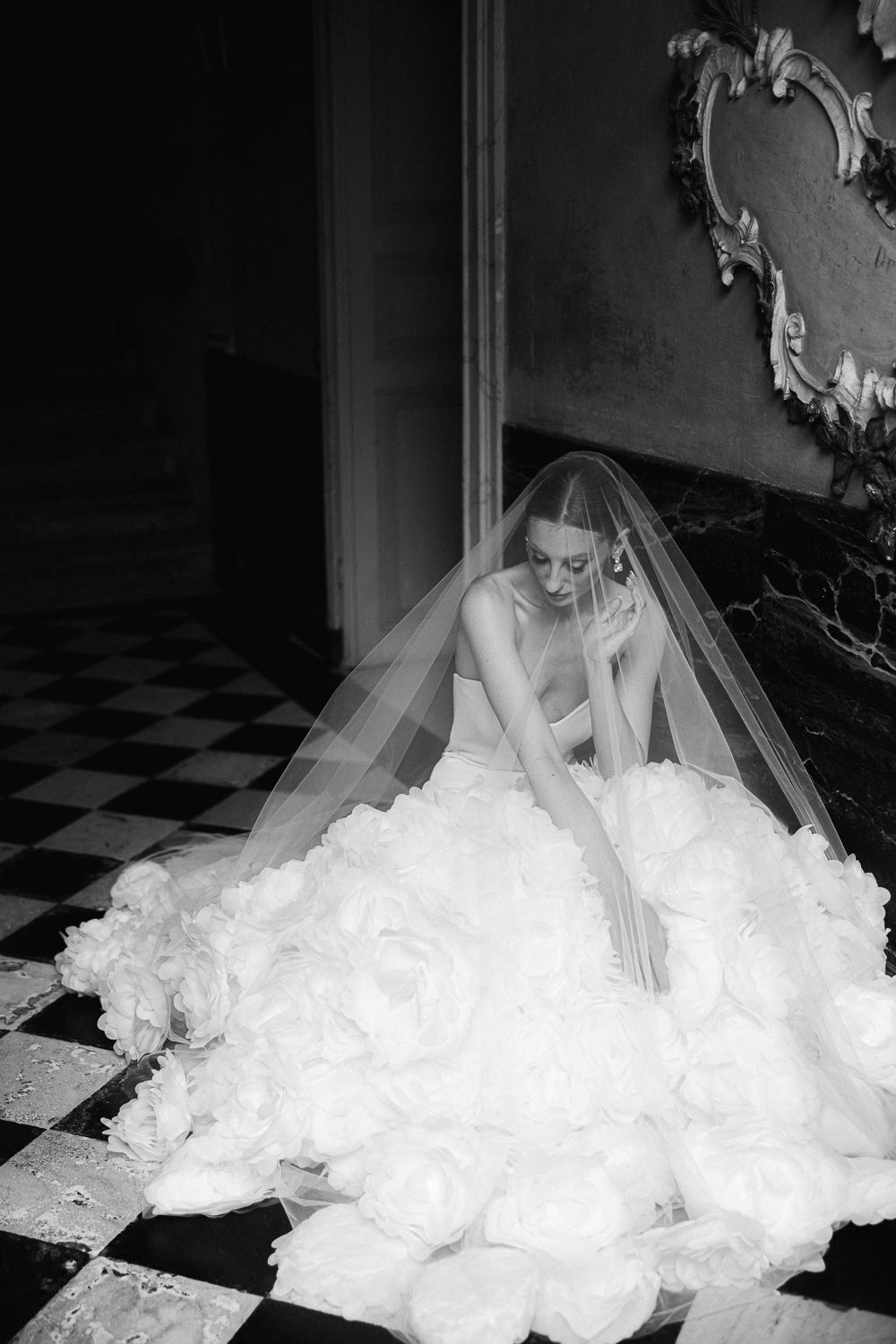 Black and white portrait of bride in ruffled ballgown seated on checkered marble floor.