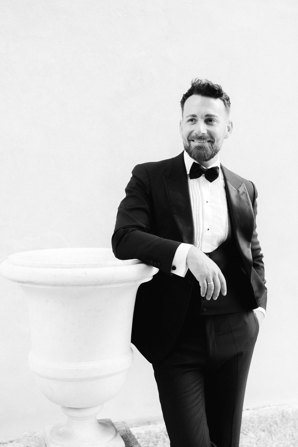 Elegant bearded groom in black tuxedo and bow tie posing beside a white stone urn in black and white photo.