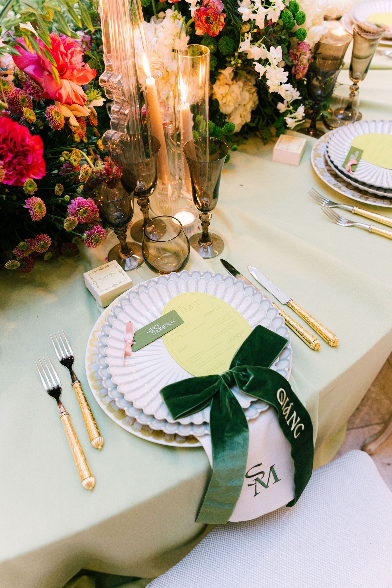 Elegant wedding table setting with gold cutlery, white scalloped plates, green velvet bow napkin ring, and lush floral centerpiece.