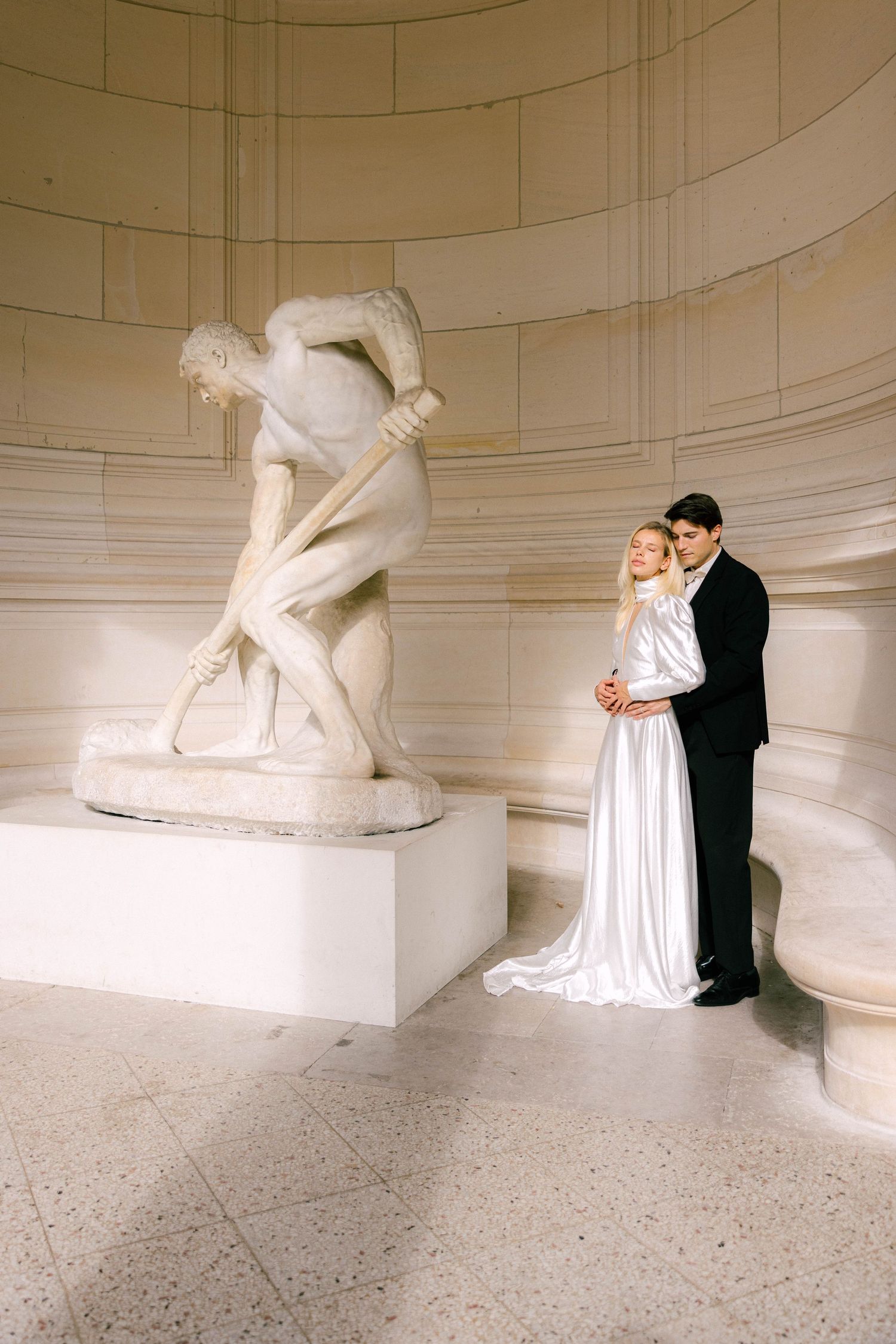 Bride in white satin gown and groom in black suit embrace beside classical marble sculpture in rotunda.