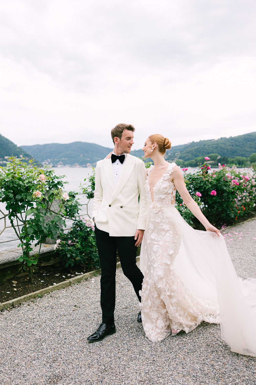 Bride in floral gown and groom in white tuxedo walking together along garden path with lake and mountains behind.