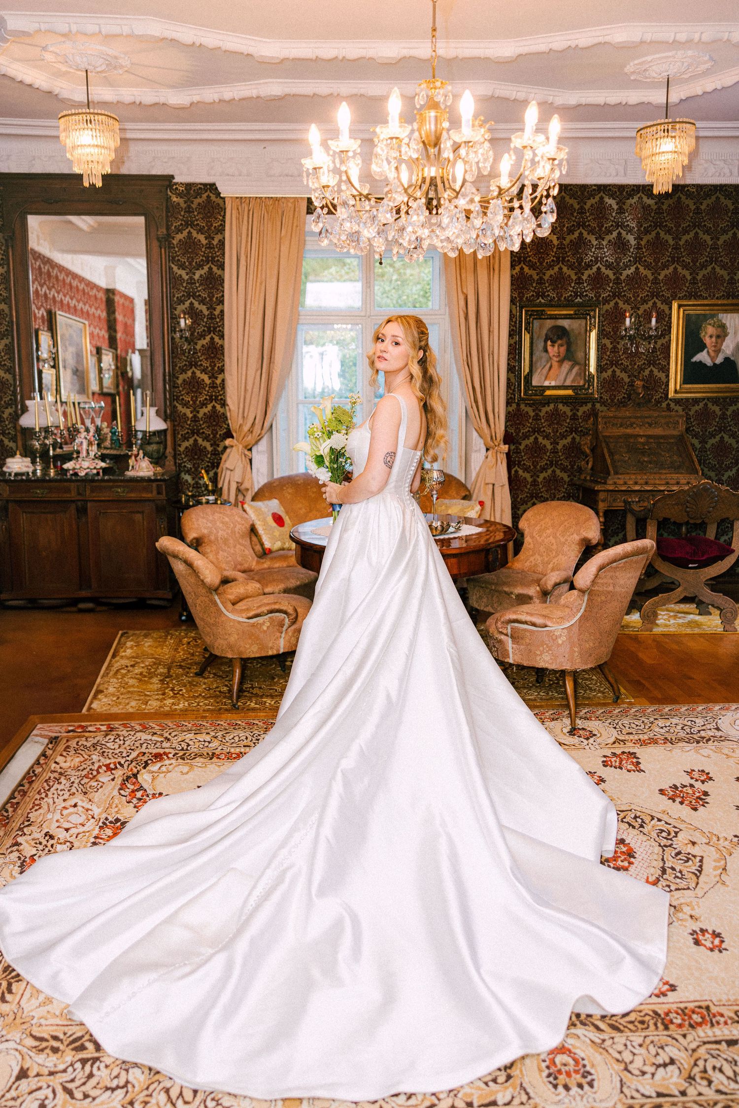 Bride in stunning white satin gown with long train standing in ornate vintage room with crystal chandelier.