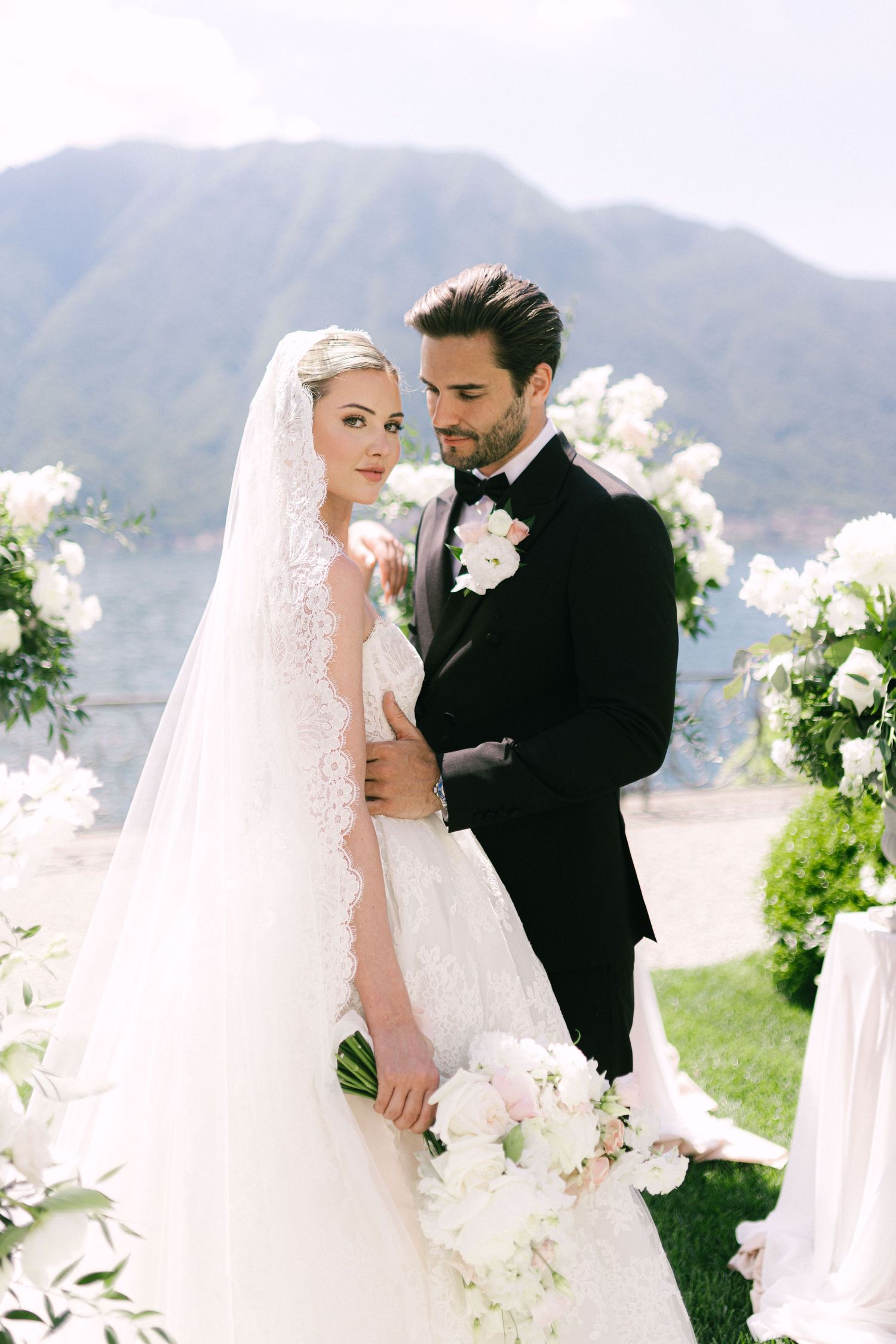 Bride in lace wedding gown with veil and groom in black tuxedo embracing at romantic lakeside ceremony.
