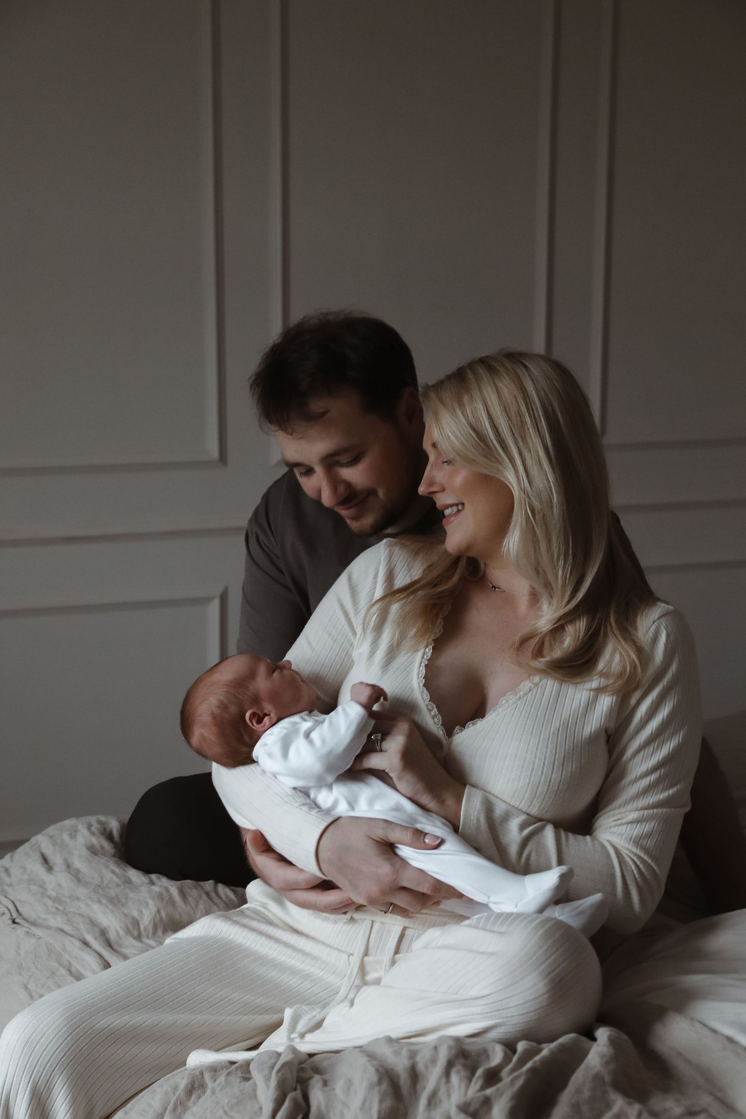 A couple sitting on bed bond with their newborn baby in a cozy, dimly lit room with white walls.