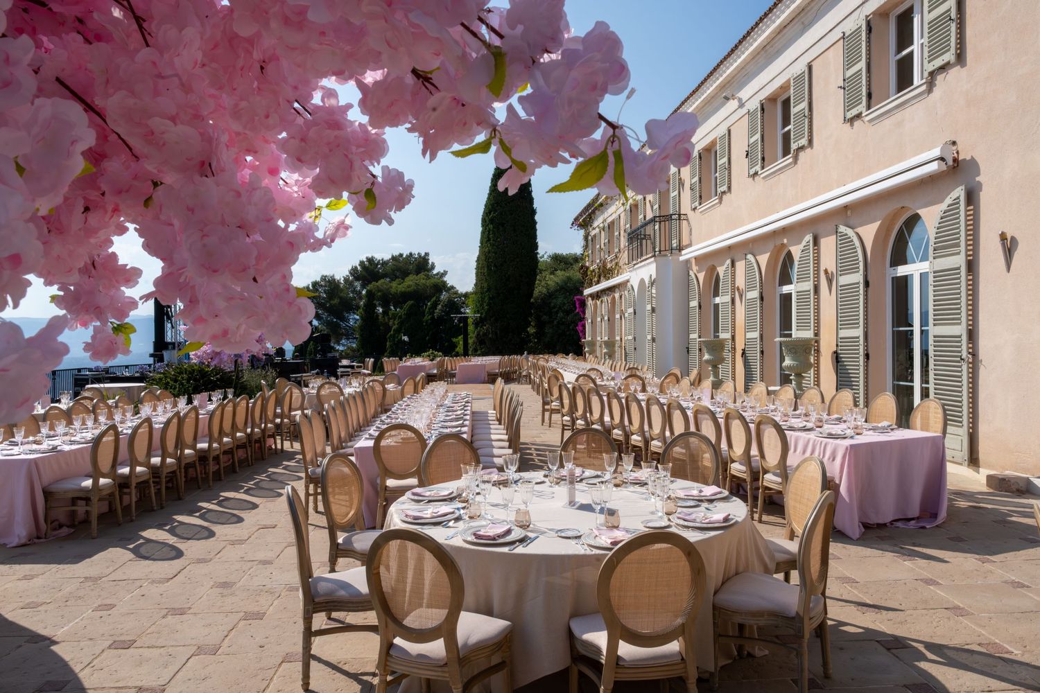 Elegant outdoor wedding reception on a terrace with round tables, gold chairs, and pink cherry blossoms.
