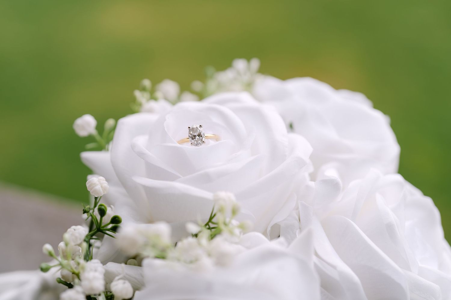 Diamond engagement ring resting on white roses in a bridal bouquet with baby's breath against green background.