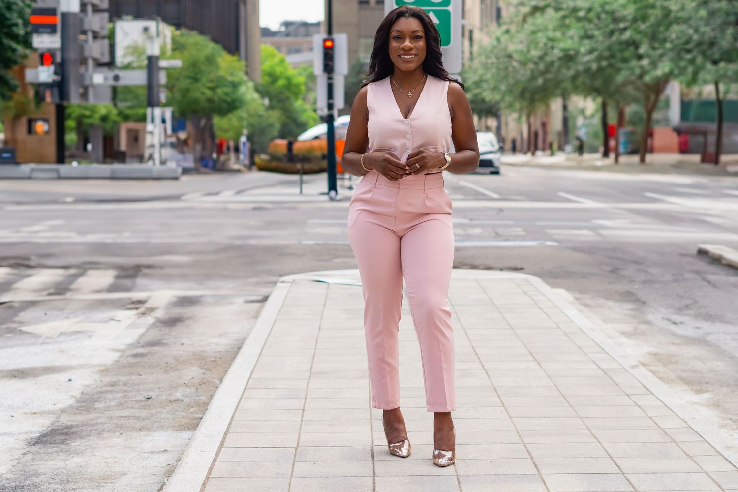 A person in a pink pantsuit and heels walks confidently down an urban sidewalk on a sunny day.
