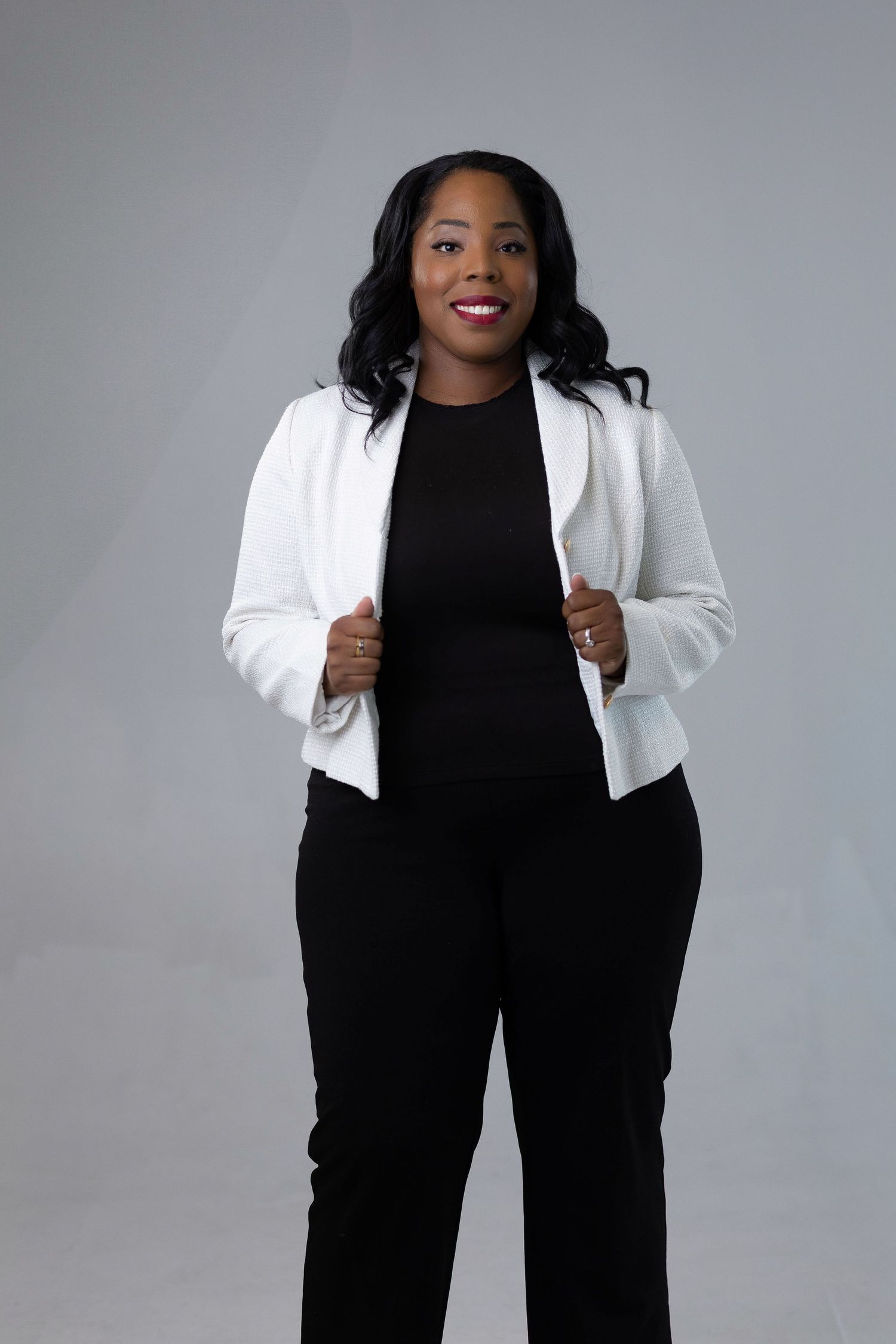 Professional portrait against gray background showing person in black outfit with crisp white blazer and confident smile.