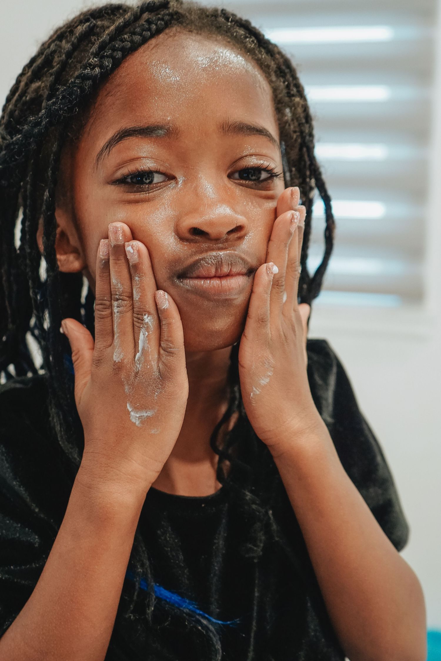 Young person with braids pressing palms against cheeks while posing indoors.