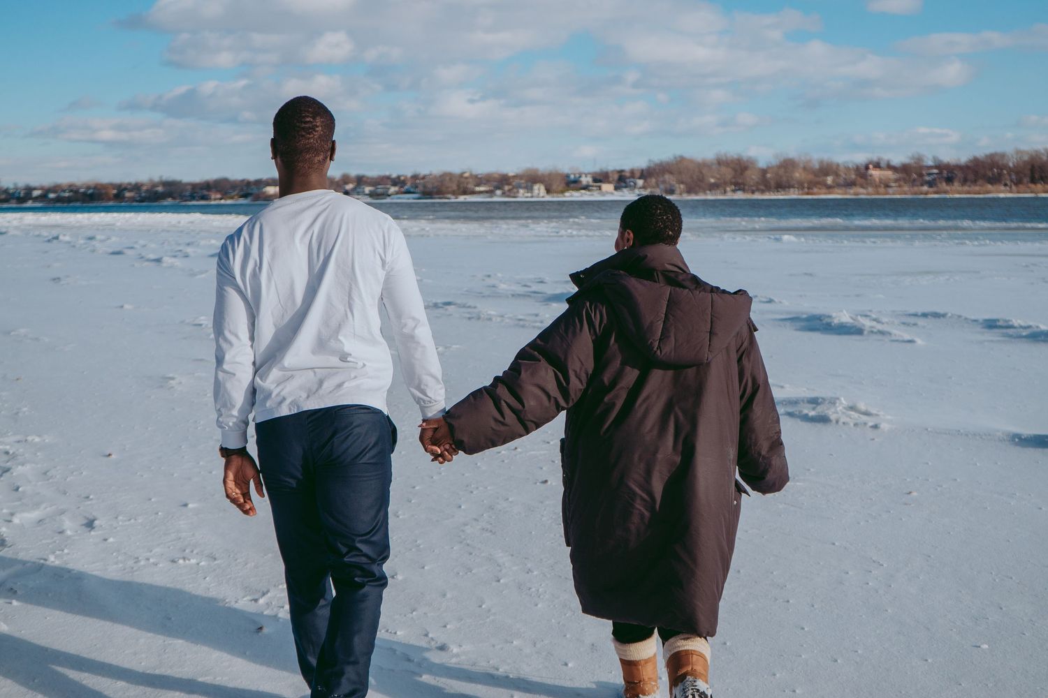 A couple walks hand in hand across a snowy winter landscape near a frozen lake.