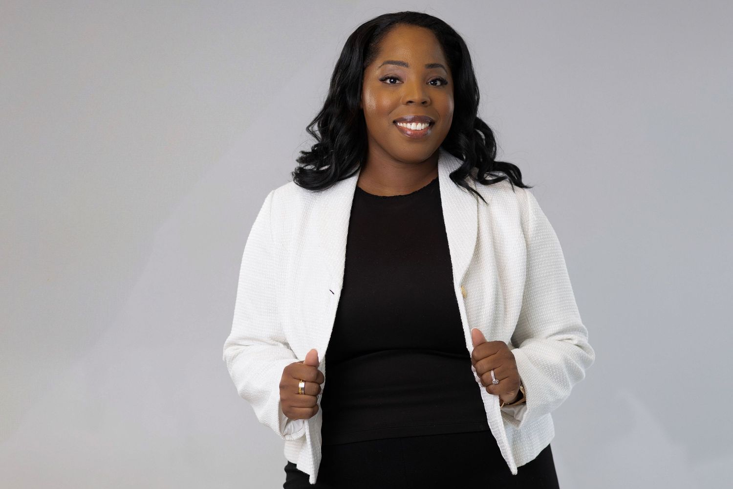 Professional portrait of a woman in a black dress and white blazer against a gray background.
