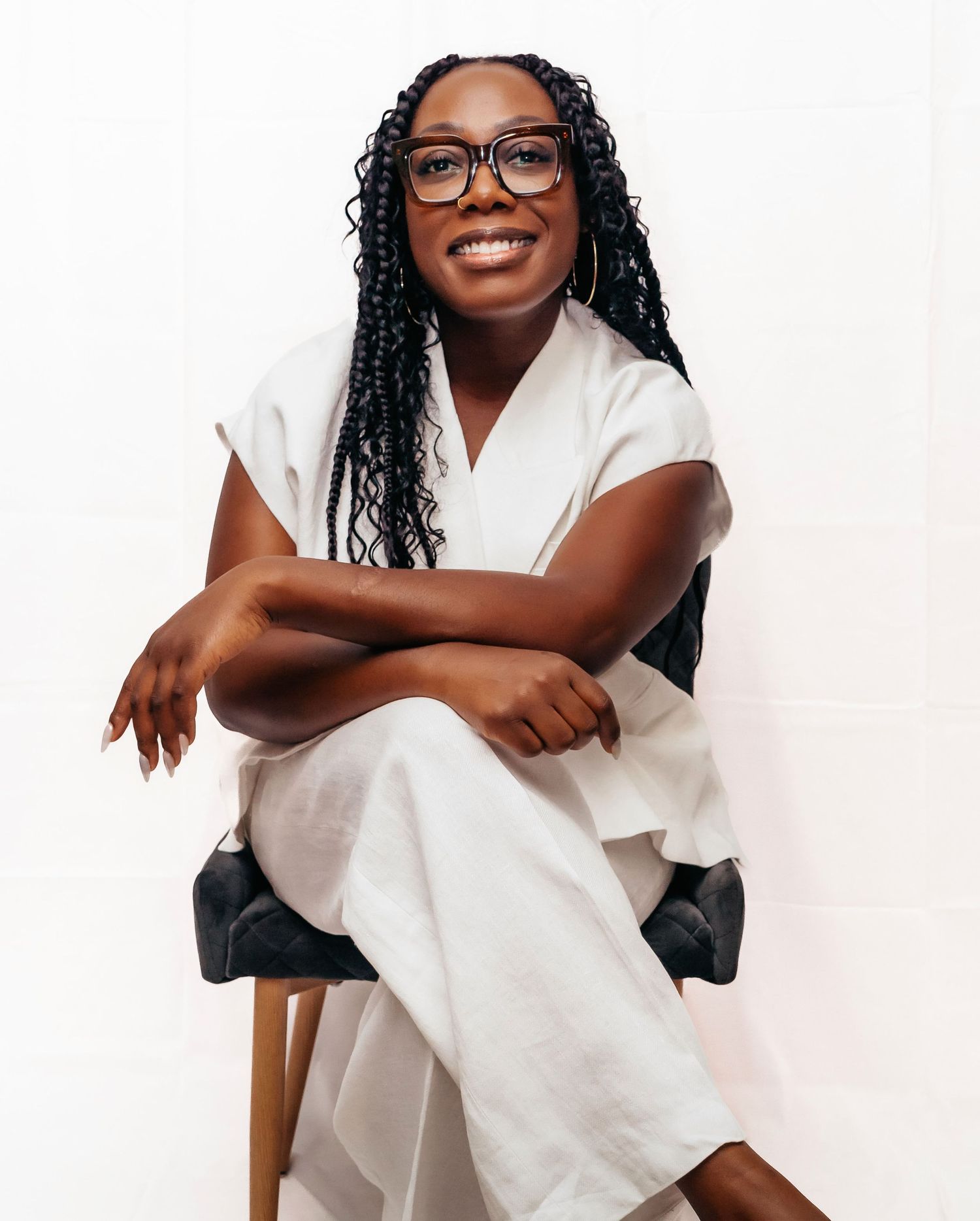 A person wearing glasses and a white outfit sits casually on a chair with crossed legs and a bright smile.