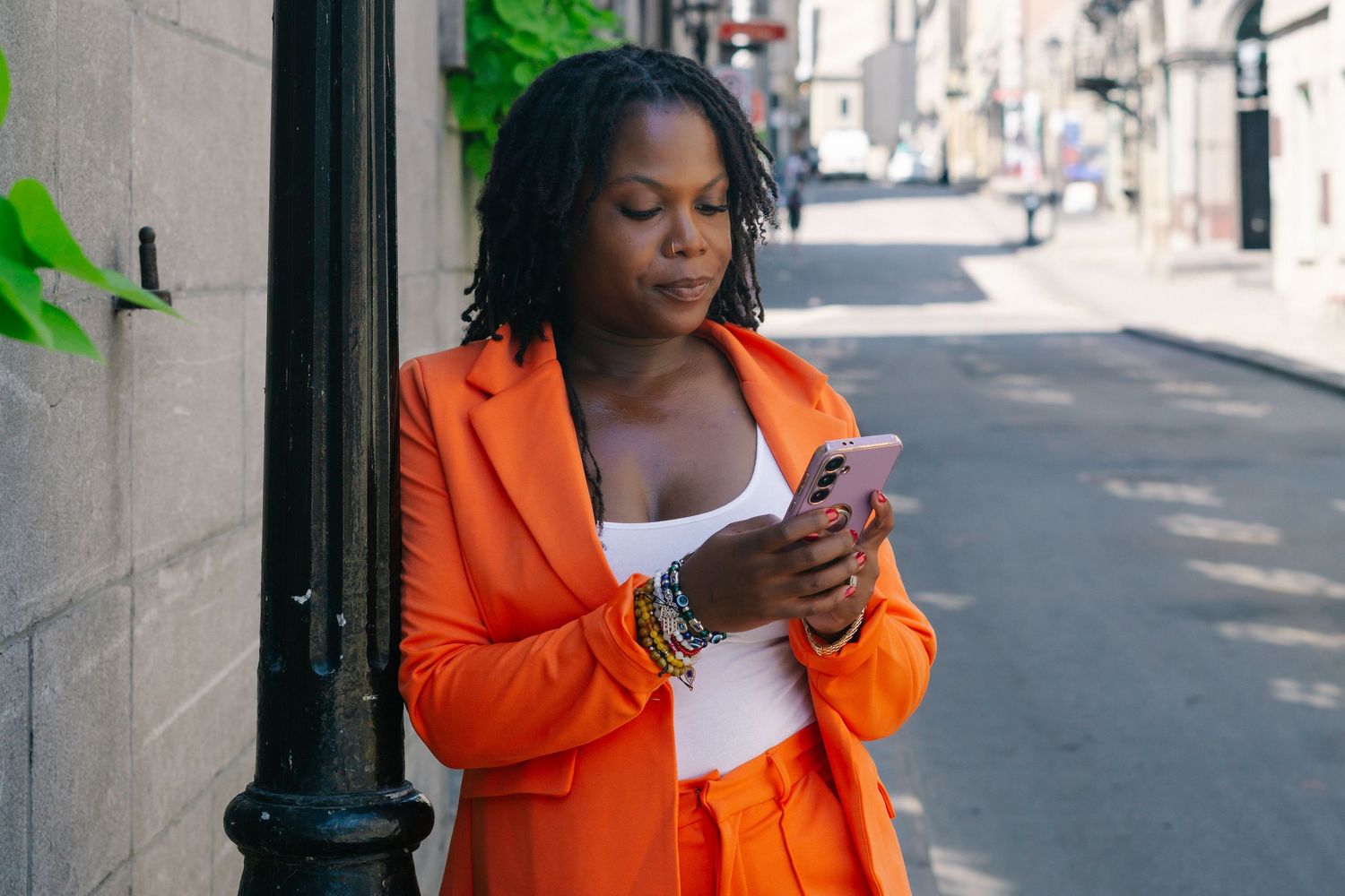 A person in a bright orange blazer stands on an urban street looking at their phone.
