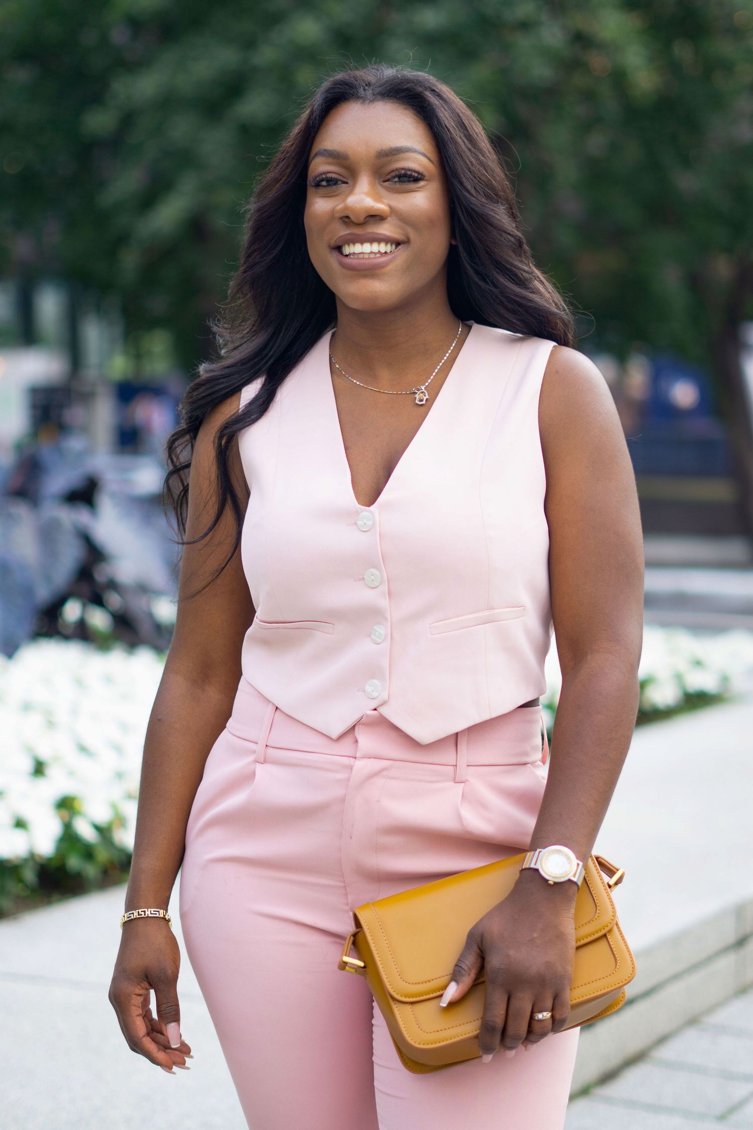 A woman in a light pink sleeveless outfit poses confidently while holding a yellow clutch purse outdoors.