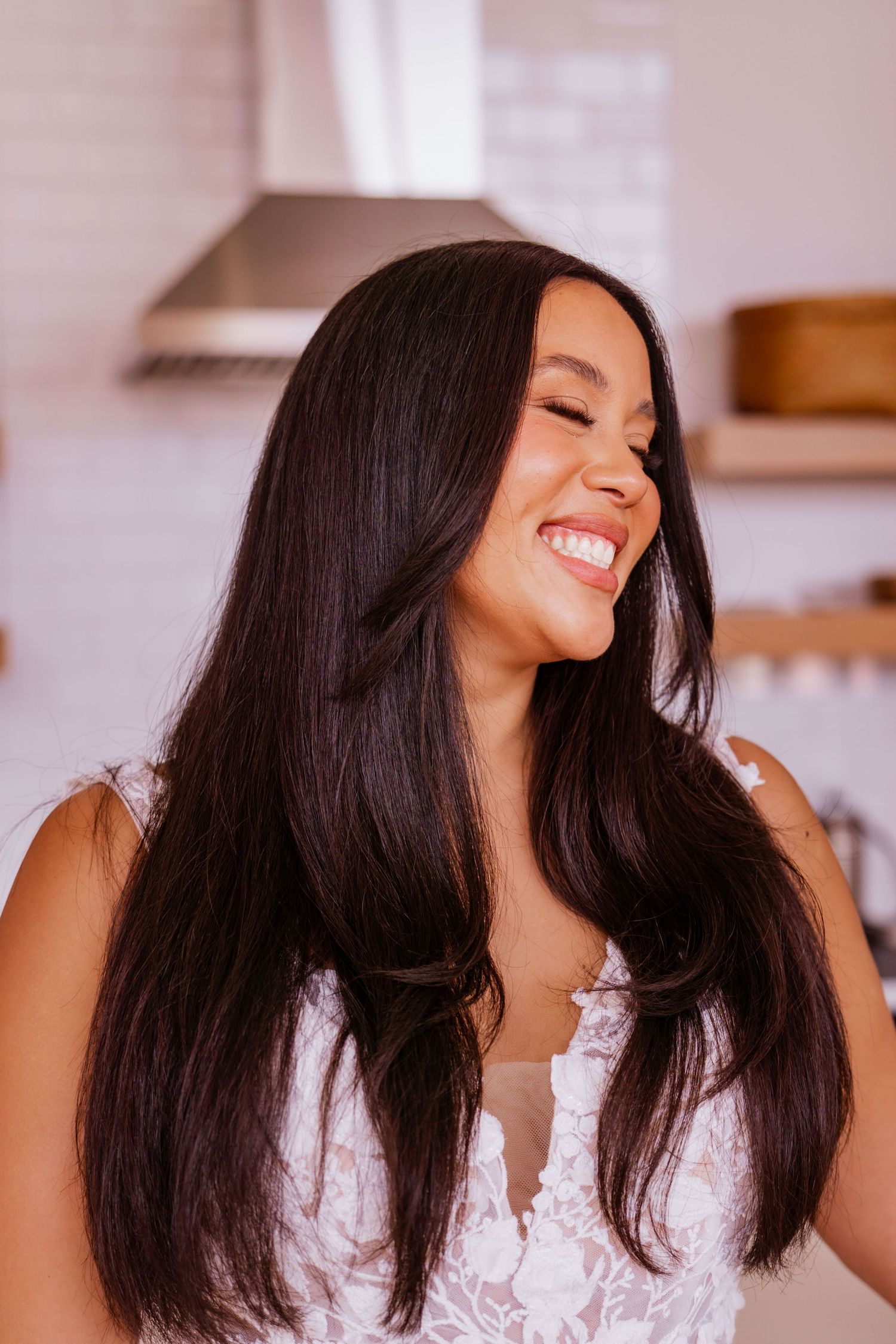 A candid moment captures someone with long dark hair and radiant smile wearing a white lace top in a kitchen setting.