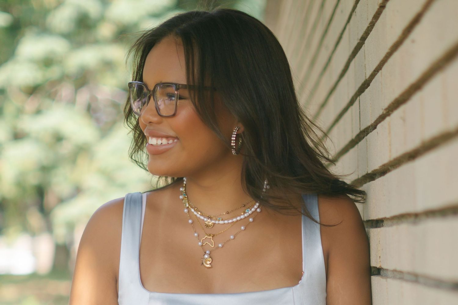 A person with glasses and dark hair smiles warmly while sitting outdoors in natural lighting.