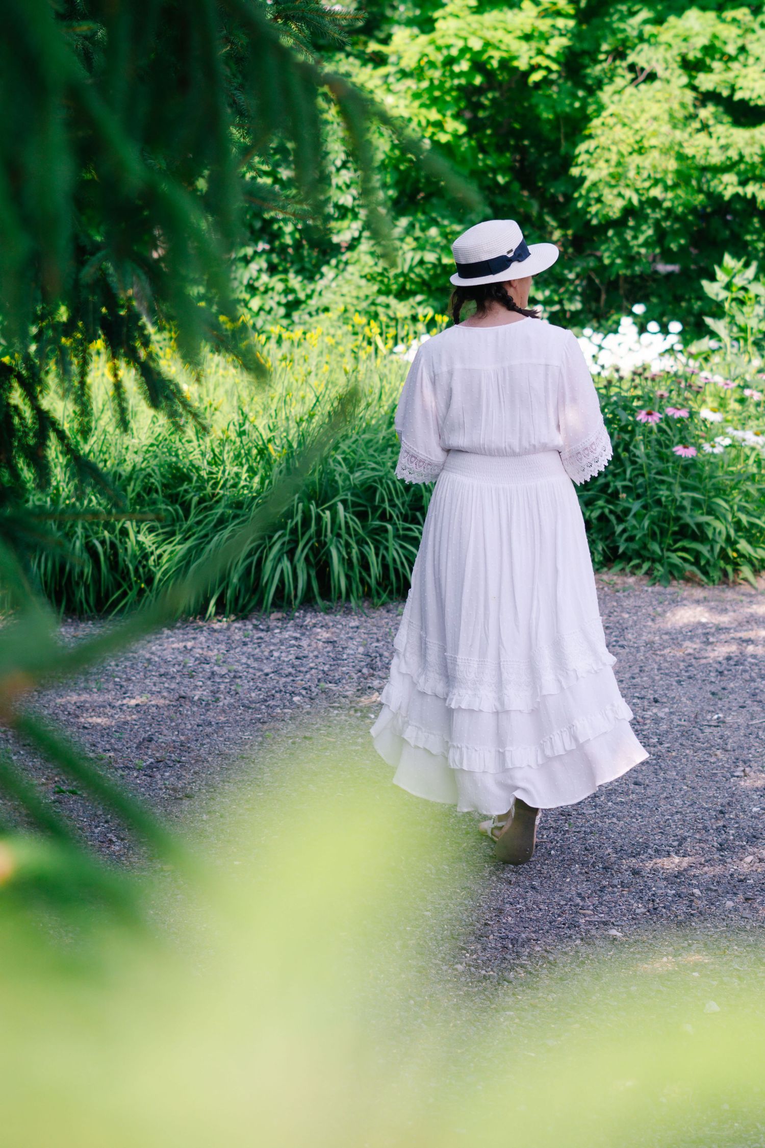 Person in flowing white dress and hat walks along garden path surrounded by lush greenery and summer foliage.