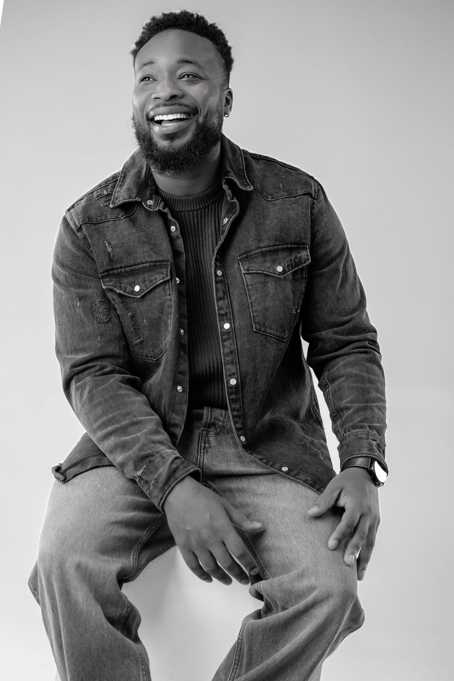 Black and white portrait of someone wearing denim jacket and jeans while sitting and smiling joyfully.