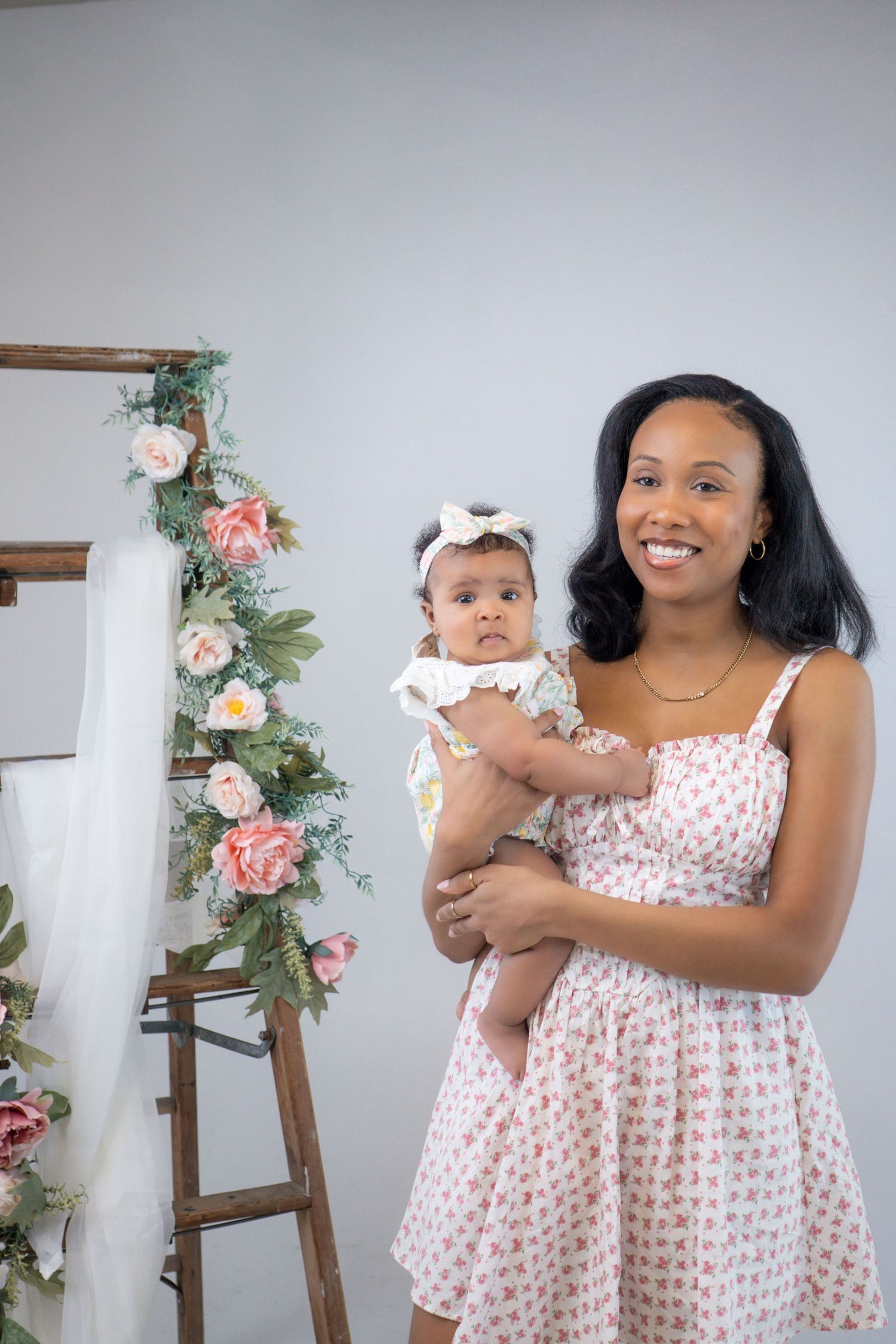 Parent in polka dot dress holds baby against backdrop with floral decorated ladder.