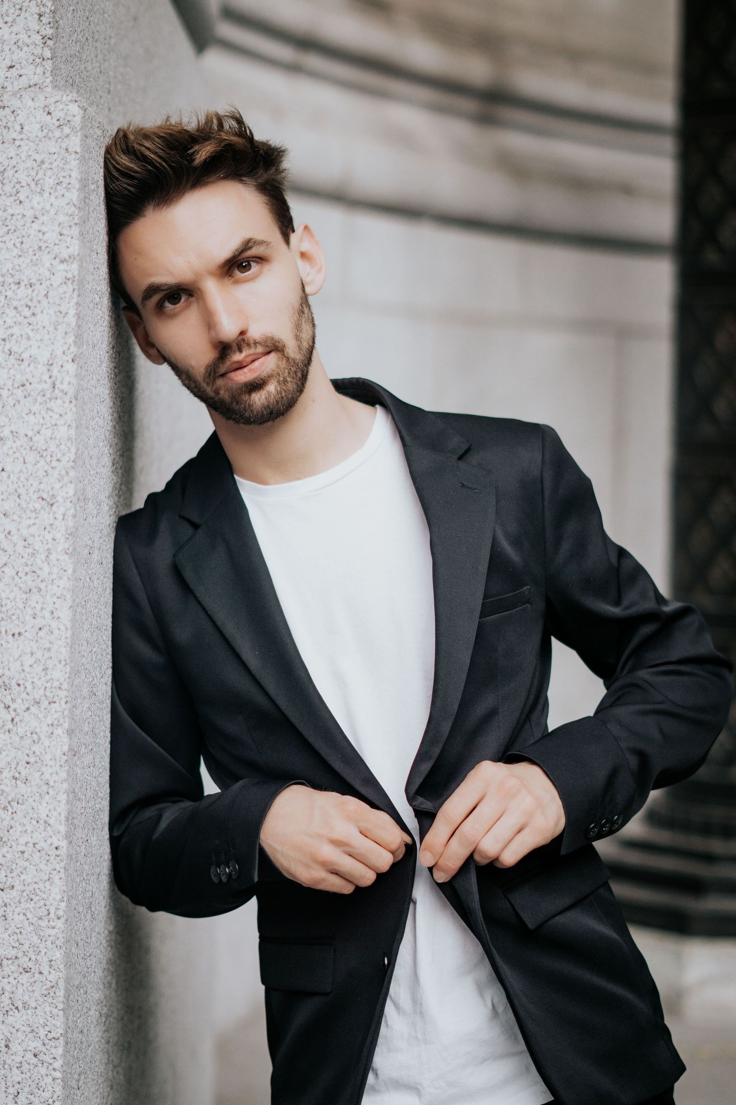 Professional wearing black blazer and white t-shirt adjusts jacket while posing against stone columns outdoors.