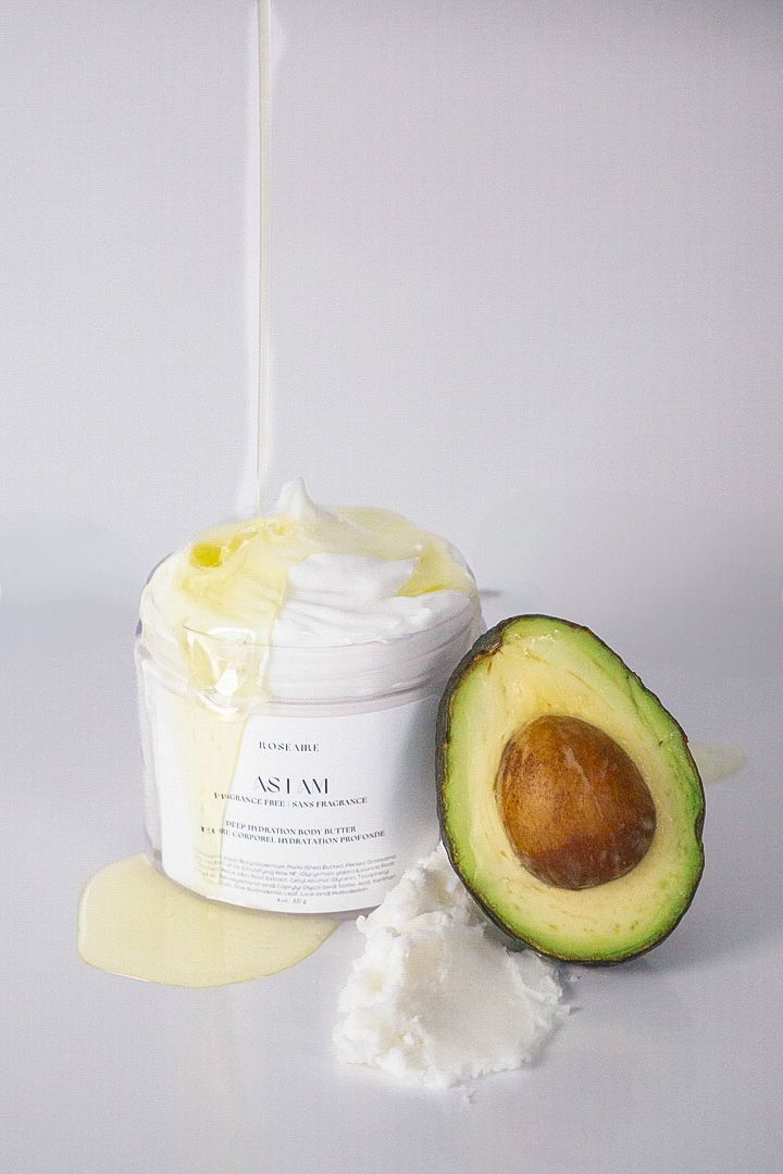White cosmetic cream jar sits beside a halved avocado on a light gray background.