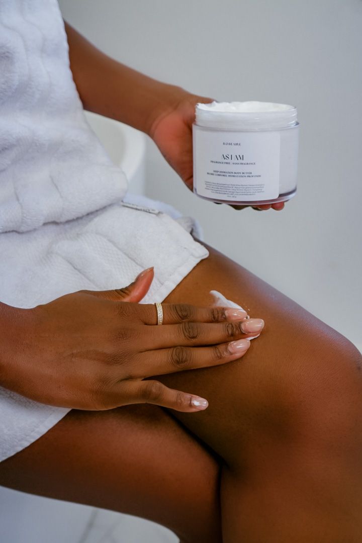 Hands applying white moisturizing cream to dark skin in a spa-like setting with white towels.