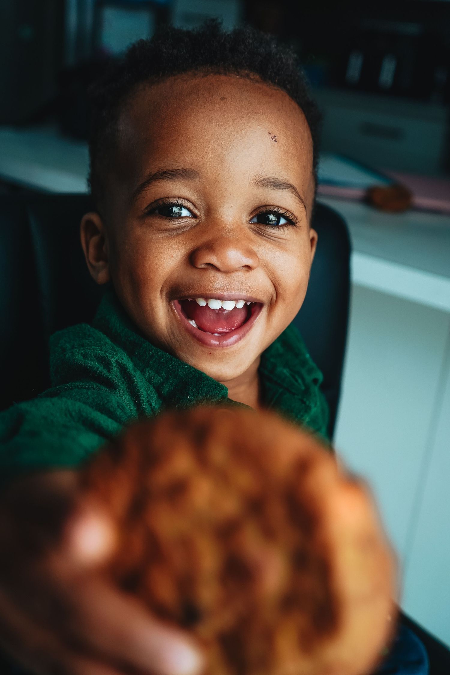Child with bright smile wearing green sweater looking at something out of frame on blurred background.
