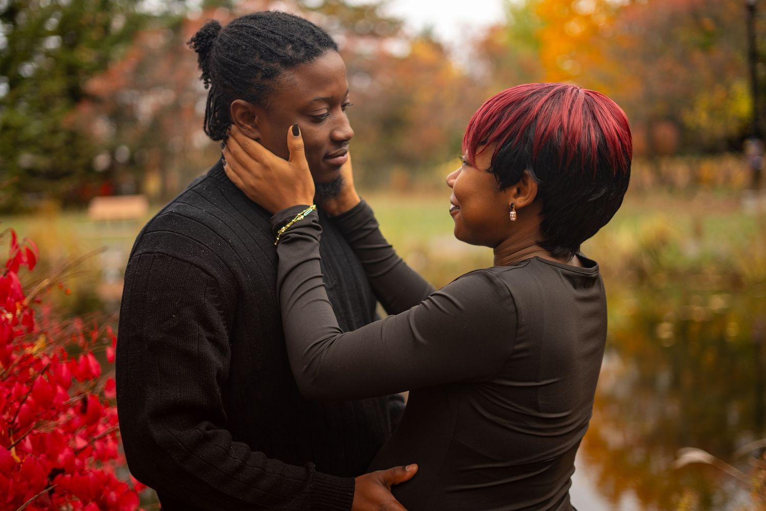 A couple embraces in front of bright red autumn bushes and golden fall trees.