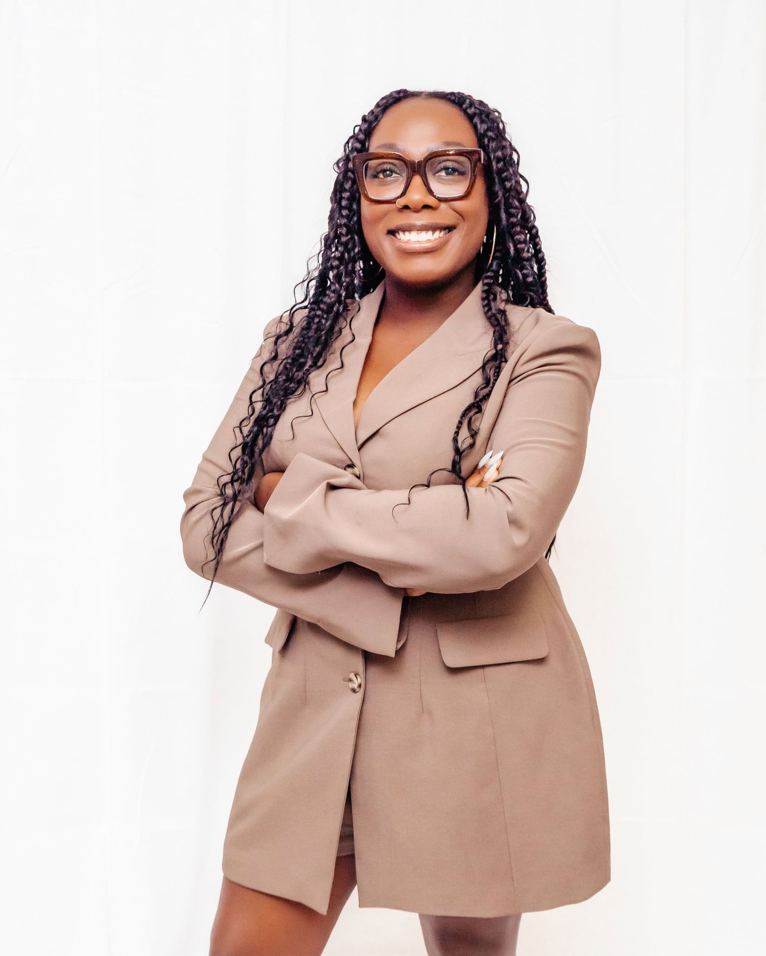 Professional wearing a tan blazer and glasses poses confidently against white background.