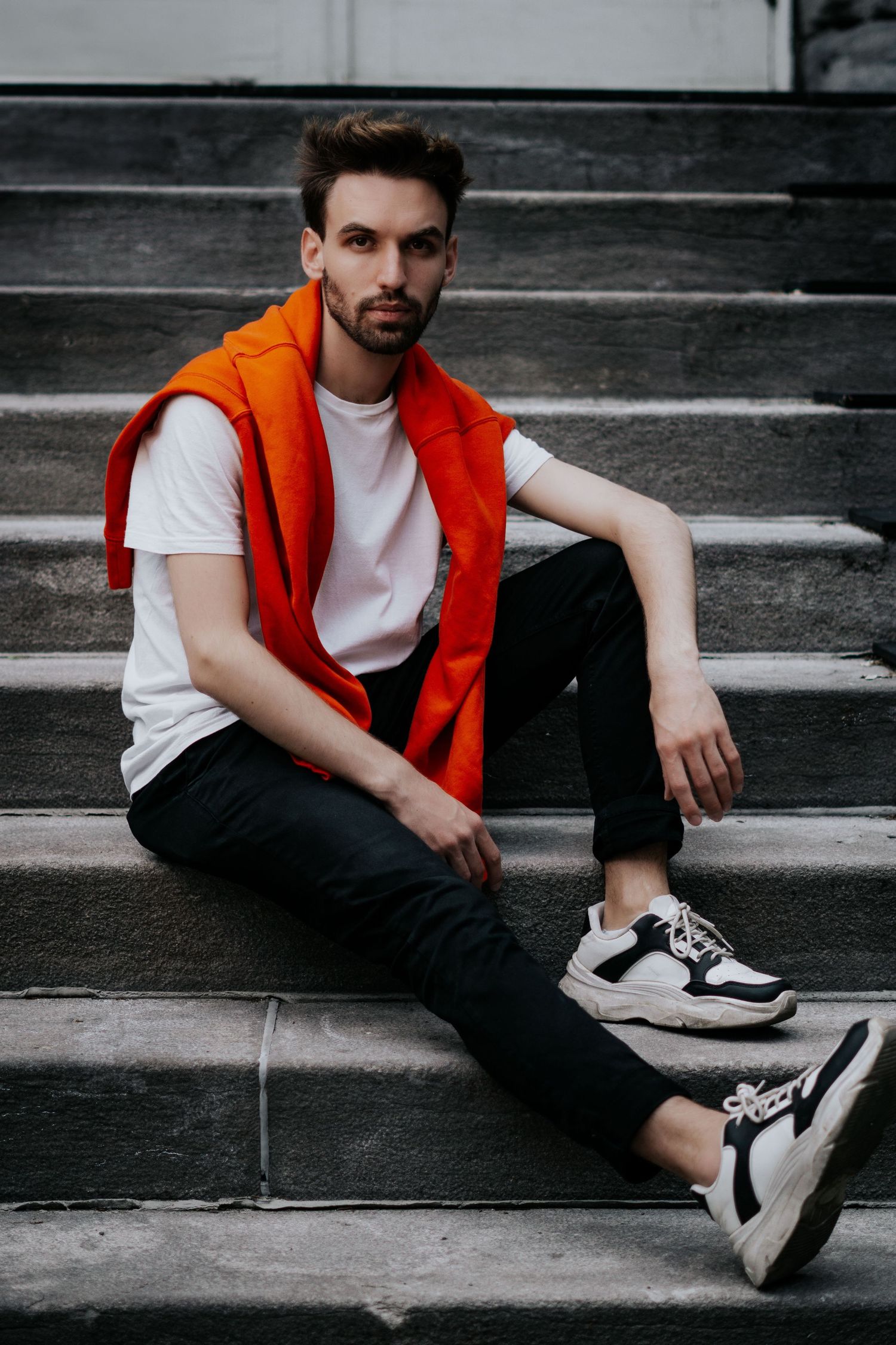 Person in orange vest and white t-shirt sits casually on concrete steps wearing black and white sneakers.