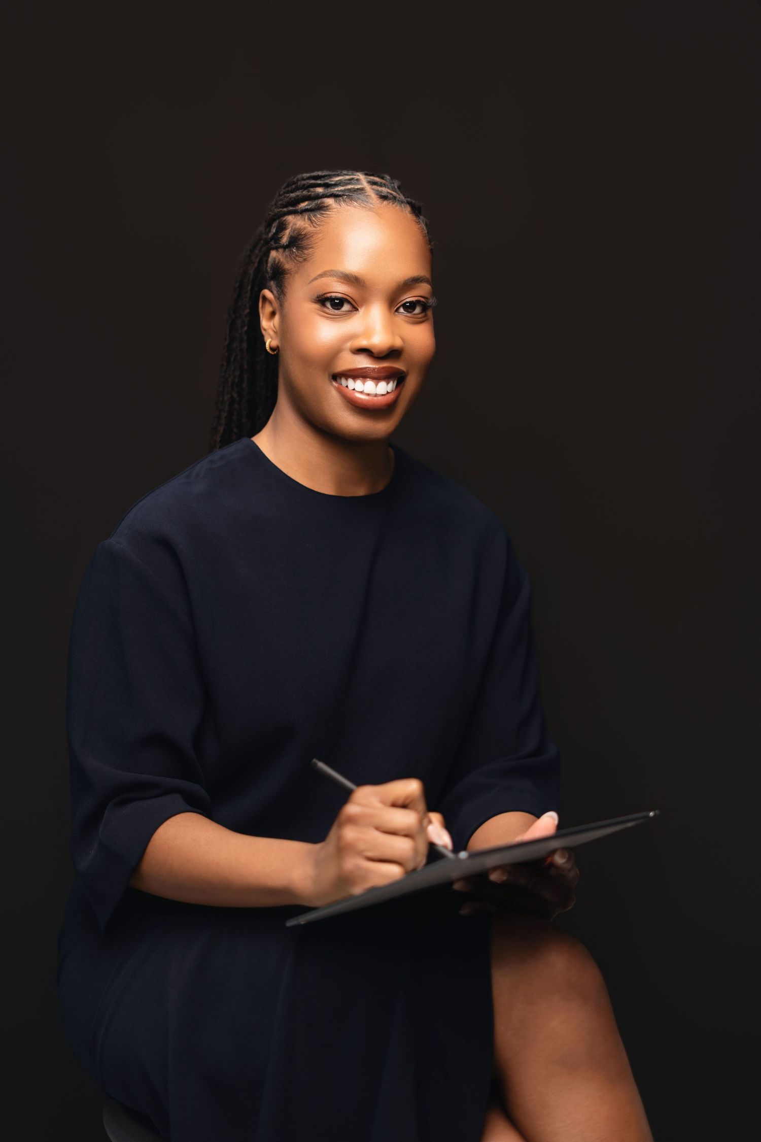 Smiling Black woman with braids holding a clipboard and pen, wearing a navy outfit against a dark background.
