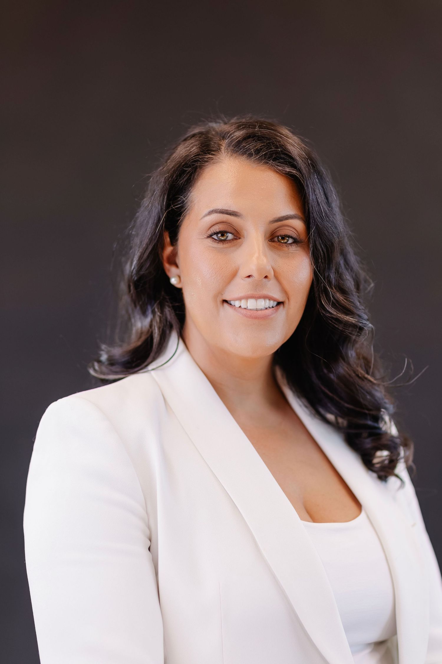 Professional headshot against dark background featuring a white blazer and confident smile.