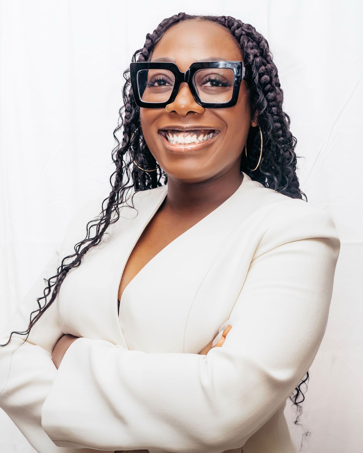 Professional portrait of someone wearing a white blazer, stylish black glasses and curly hair against a light background.