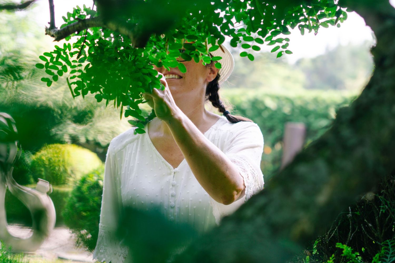 A dreamy summer scene through green foliage shows someone in a white dress standing in soft natural light.