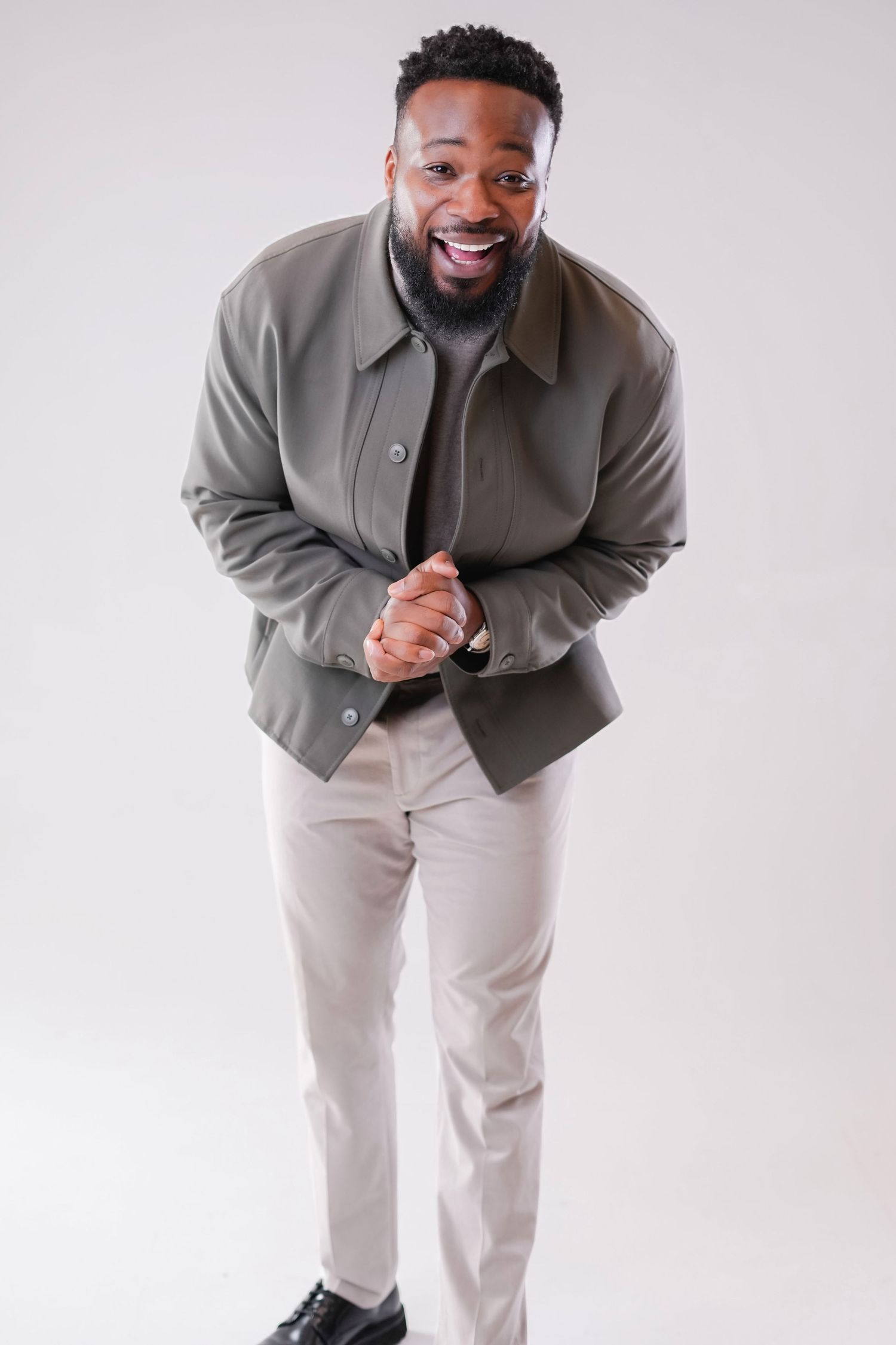 Smiling person wearing a gray jacket and khaki pants poses casually against white background.