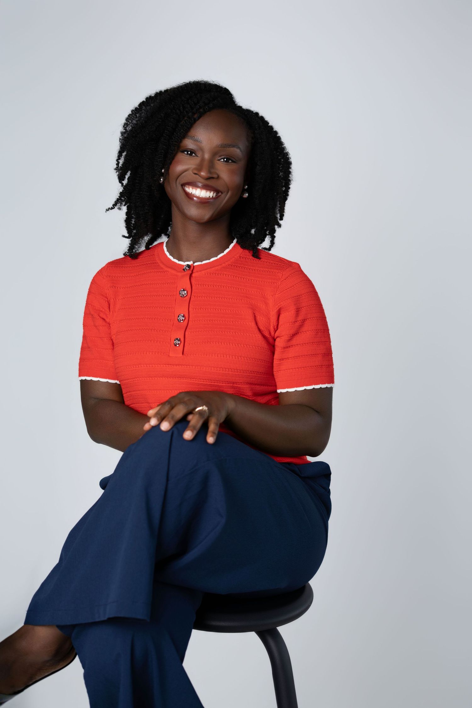 Smiling Black woman in red top and navy pants sitting on stool against gray background.