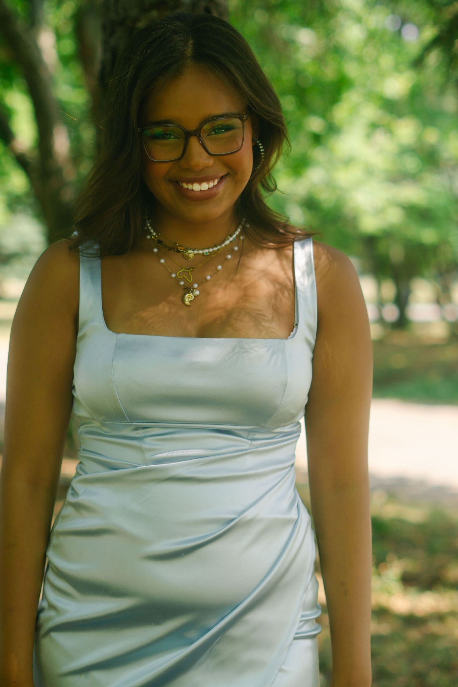 A woman in a light blue satin dress smiles while posing in an outdoor setting with trees in the background.