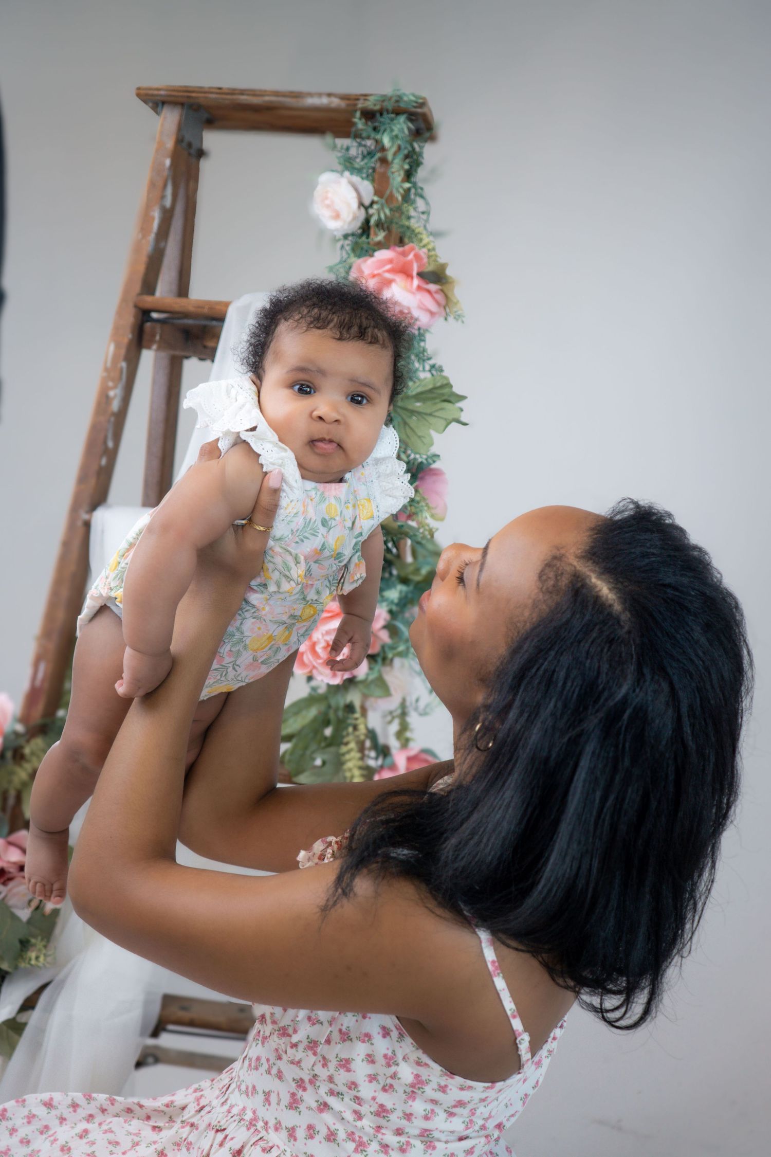 Mother lifts baby playfully in white studio setting with decorative wooden ladder and flowers.