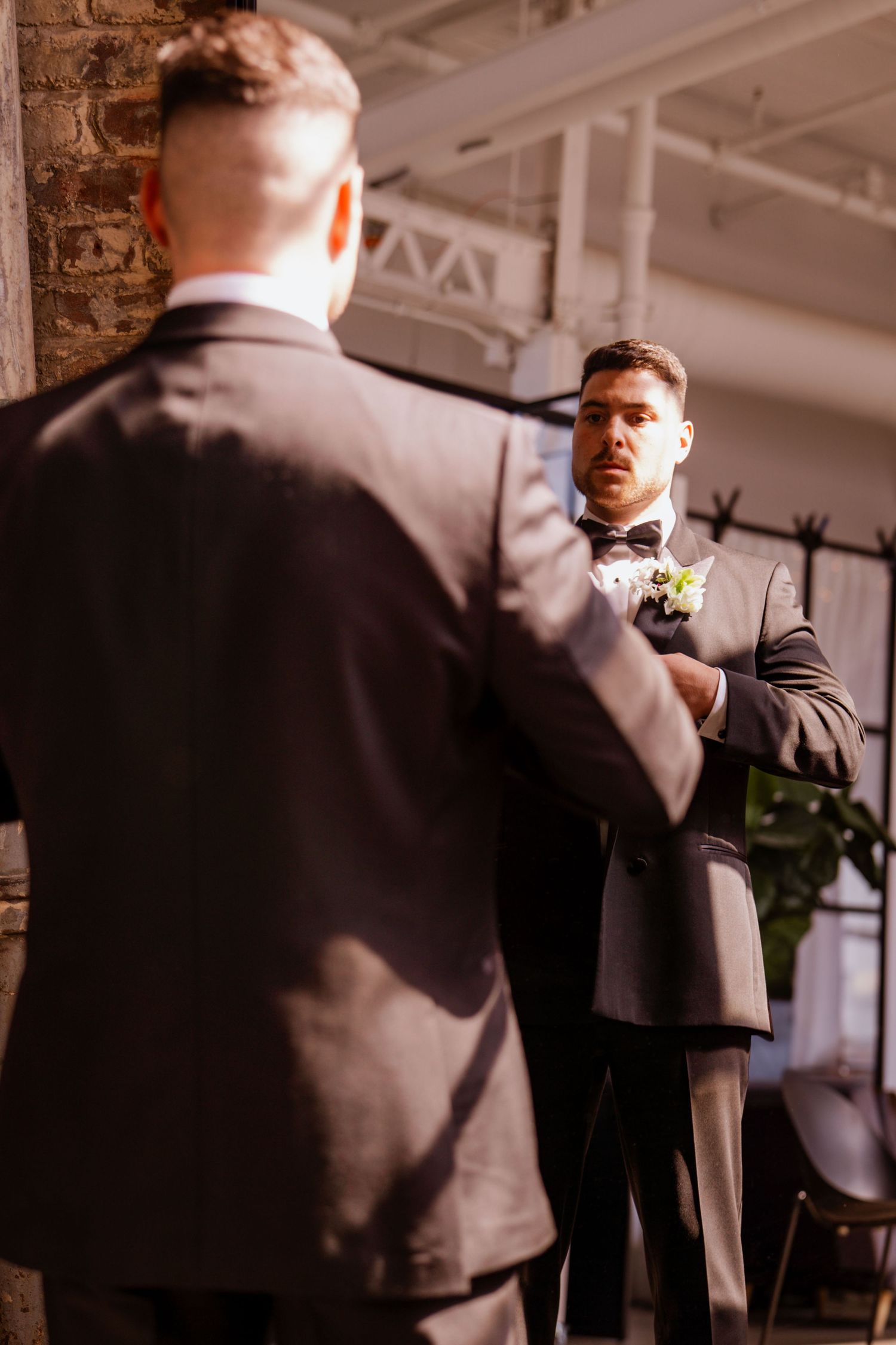 Groom in a dark suit and bow tie adjusts his outfit while looking into a full-length mirror in a rustic venue.