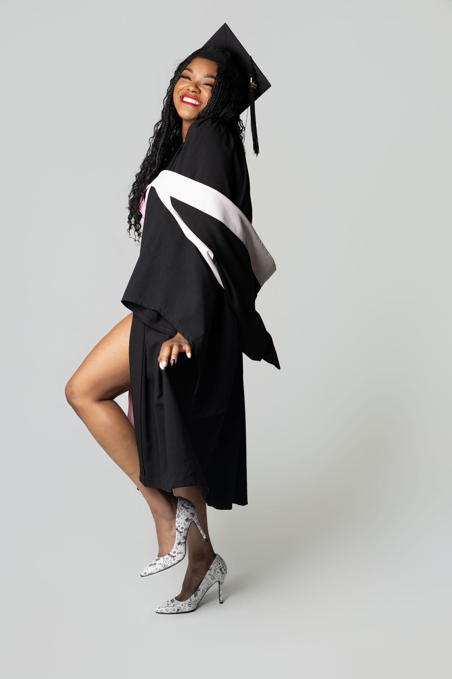 Smiling graduate woman in black cap and gown posing confidently in heels against a gray background.