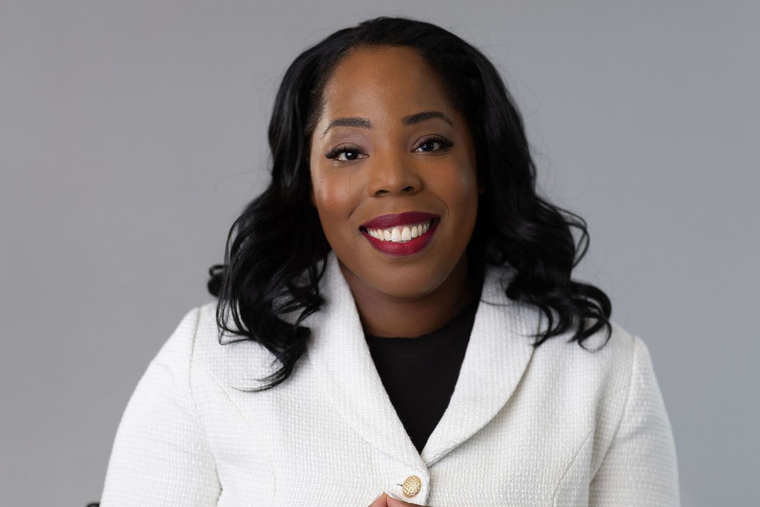 Professional headshot of woman in white blazer against gray background.