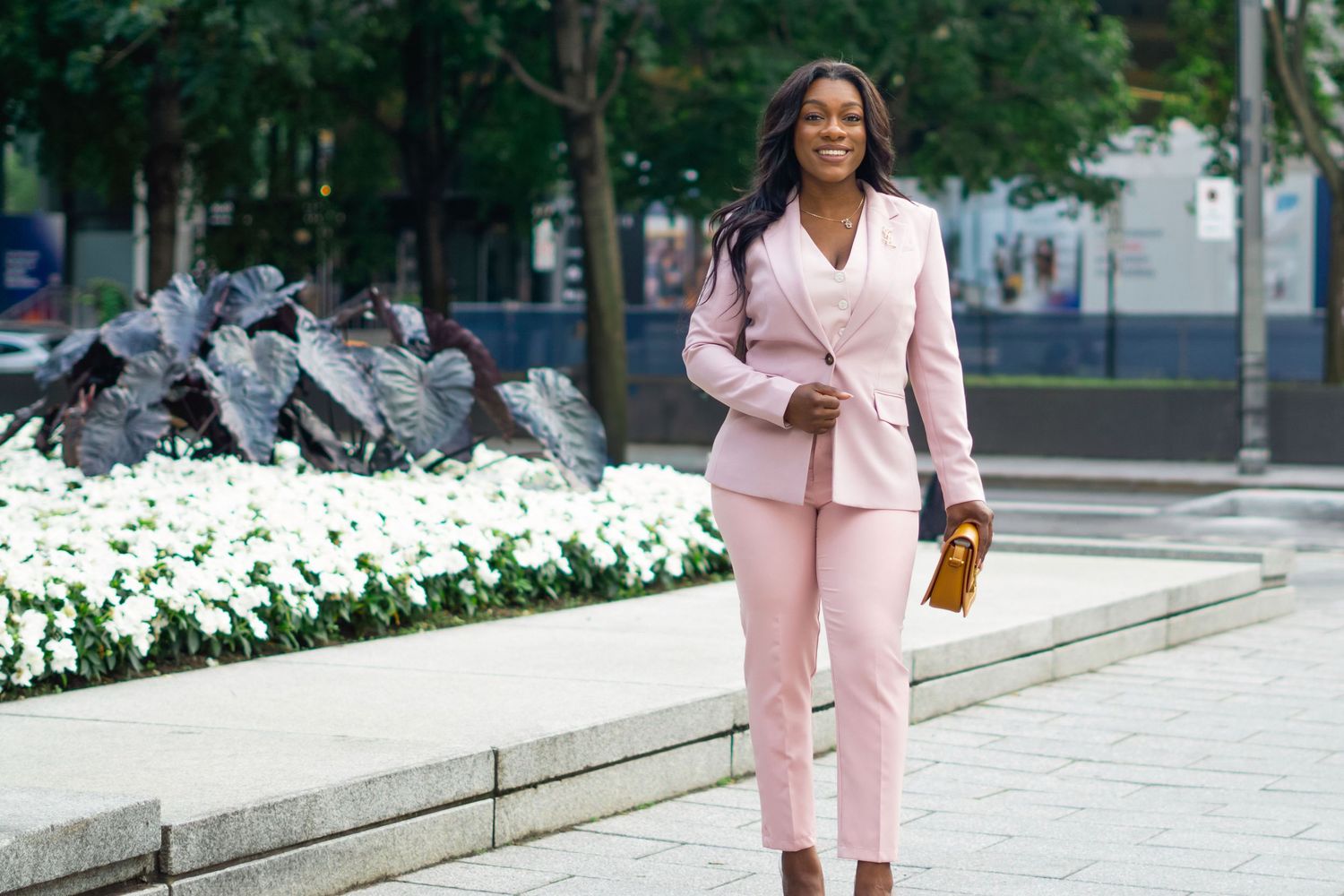 A person wearing a light pink blazer suit ensemble walks confidently on a city sidewalk near white flowers.