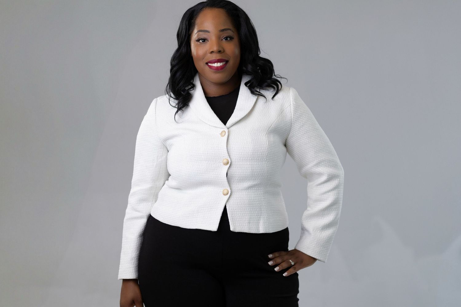 Woman in white cardigan and black dress poses confidently against gray backdrop.