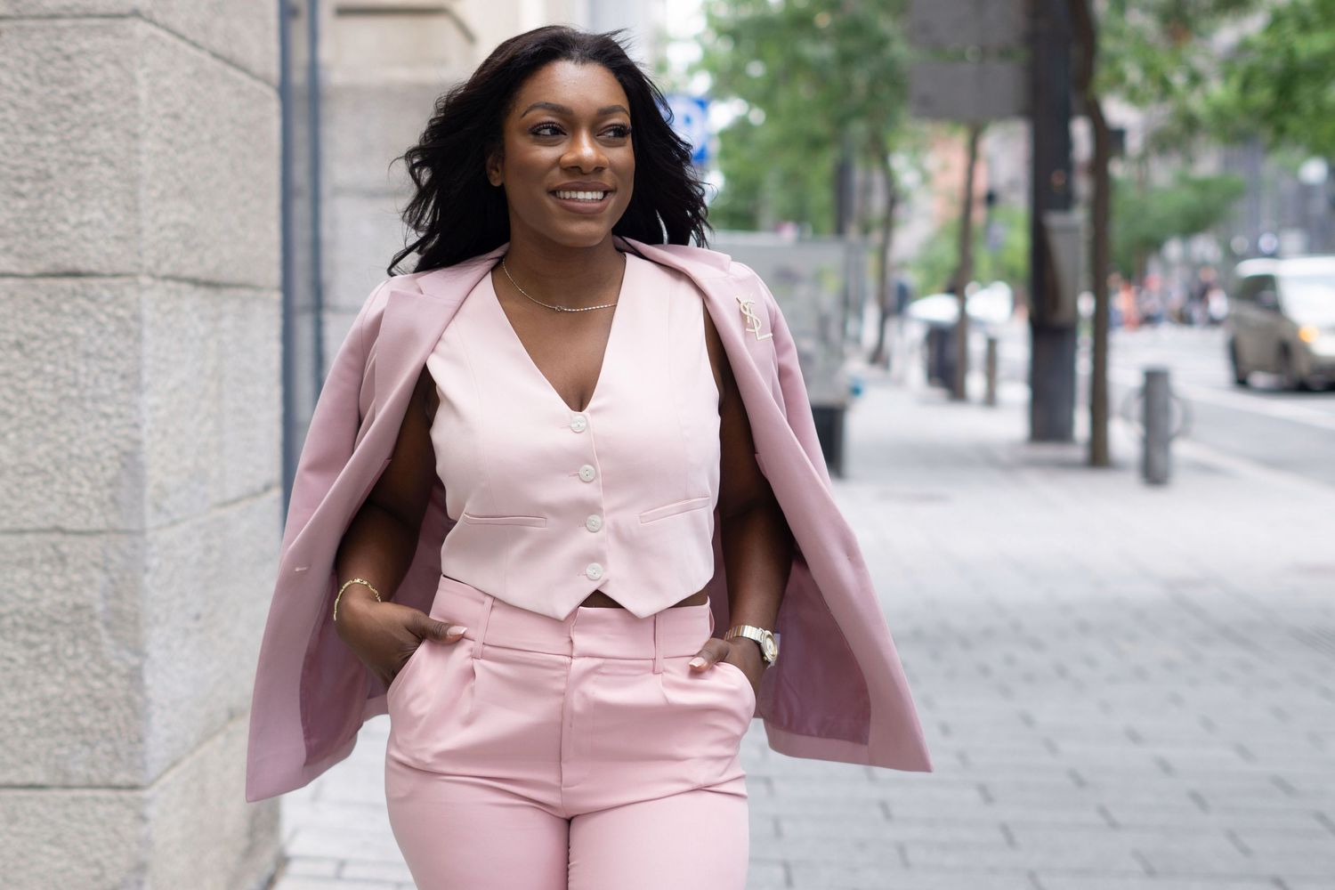 A person wearing a stylish pink pantsuit with a vest walks confidently down an urban sidewalk.